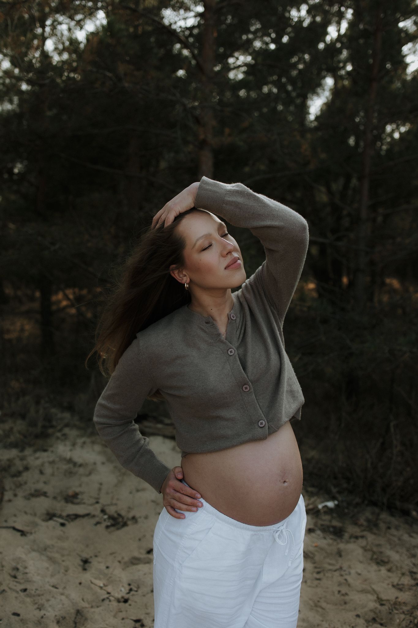 Pregnant woman in grey cardigan posing with hand in hair before pine forest.