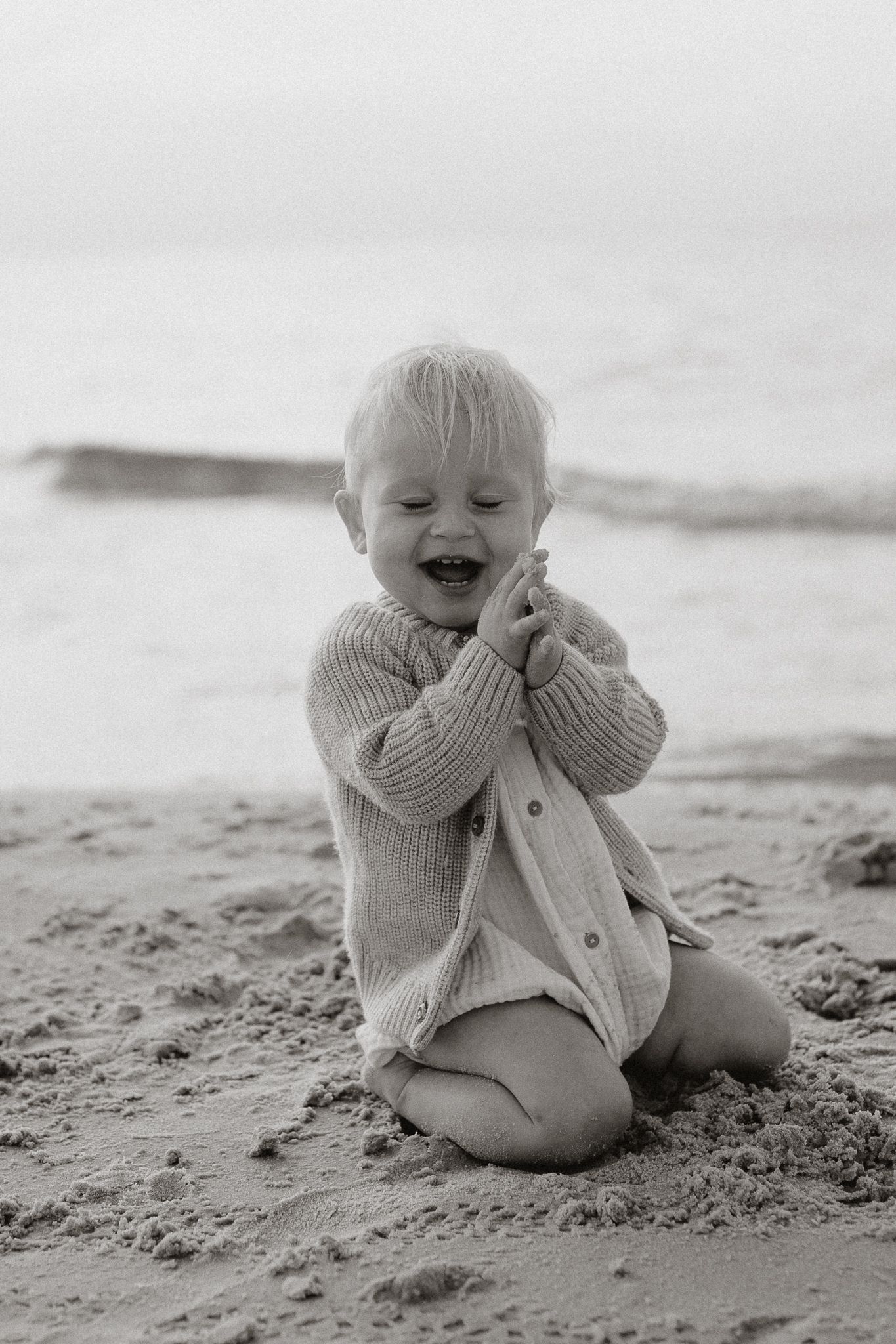 Happy toddler laughing and playing in sand on beach in black and white photo.