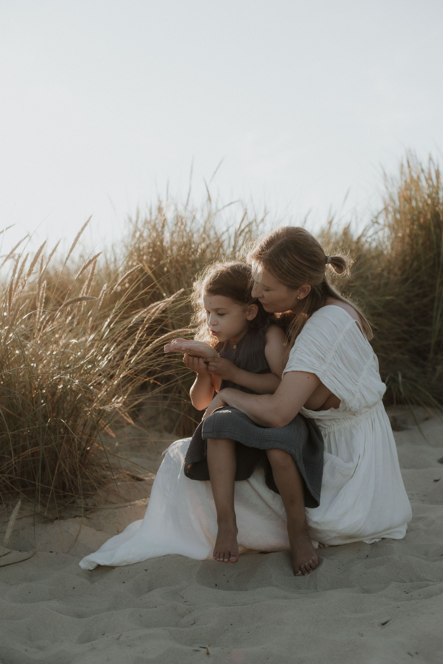 Mother in white dress kisses young child while sitting on sandy beach dunes.