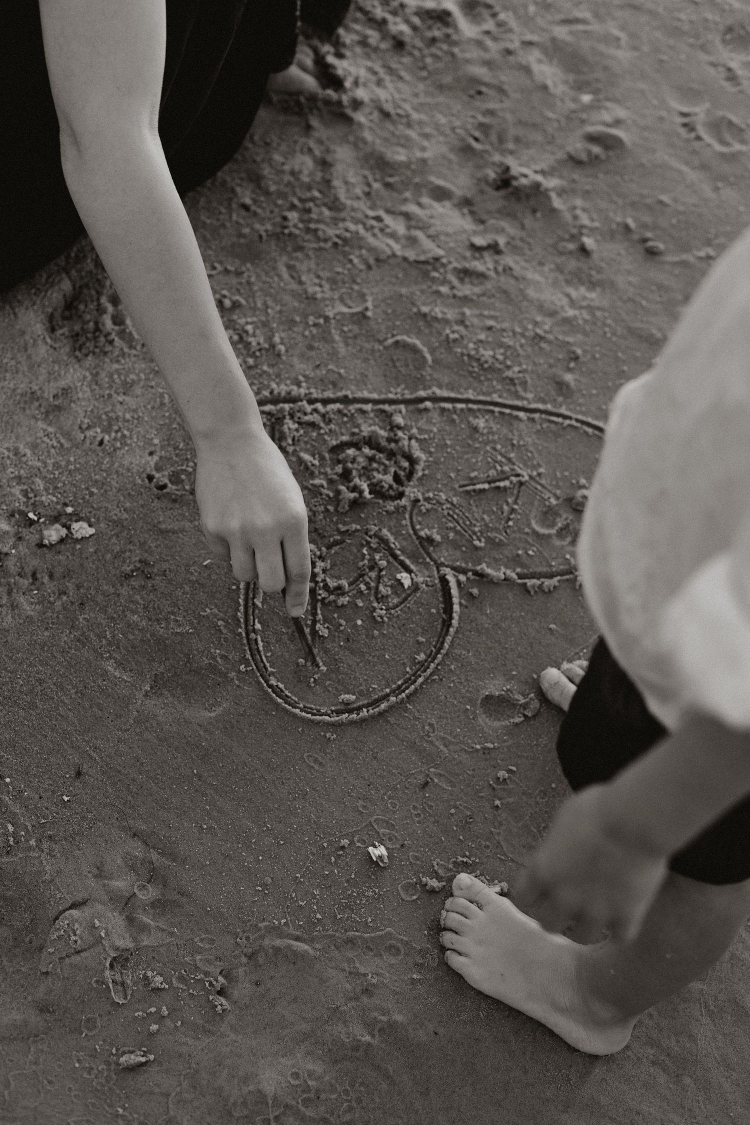 Black and white photo of person drawing in sand at beach with bare feet.