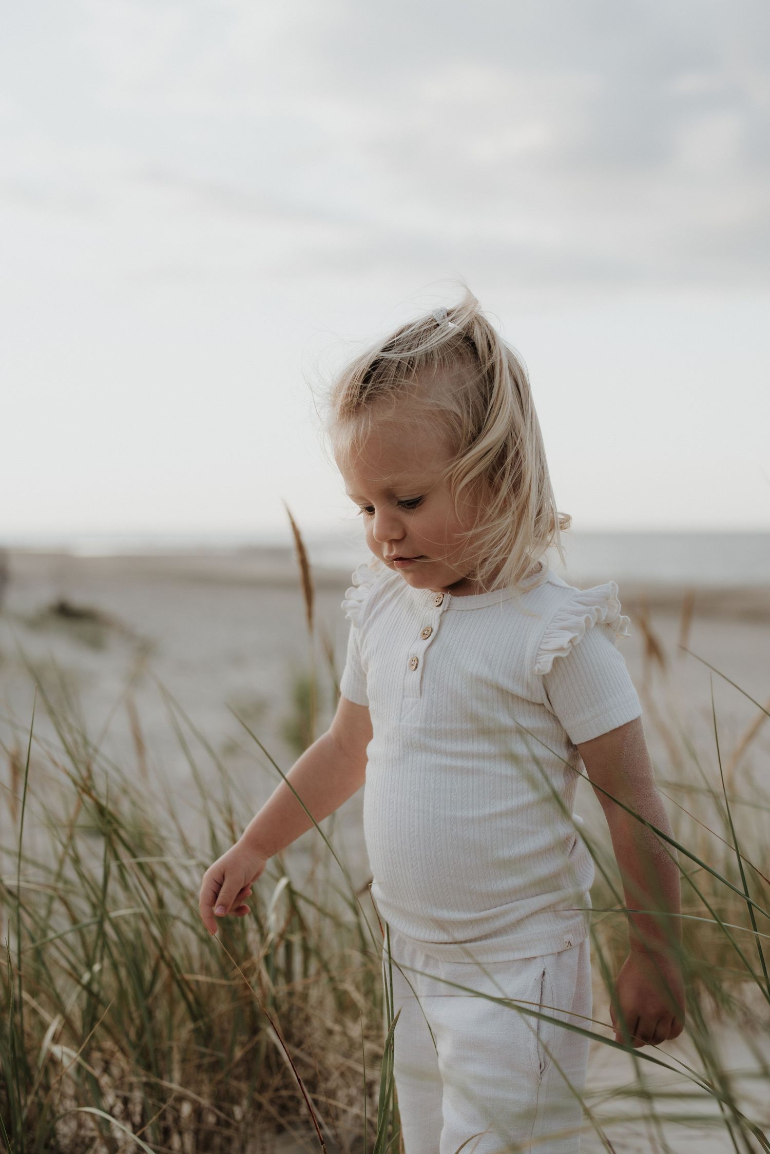Young blonde girl in white outfit exploring tall beach grass on sandy dunes.