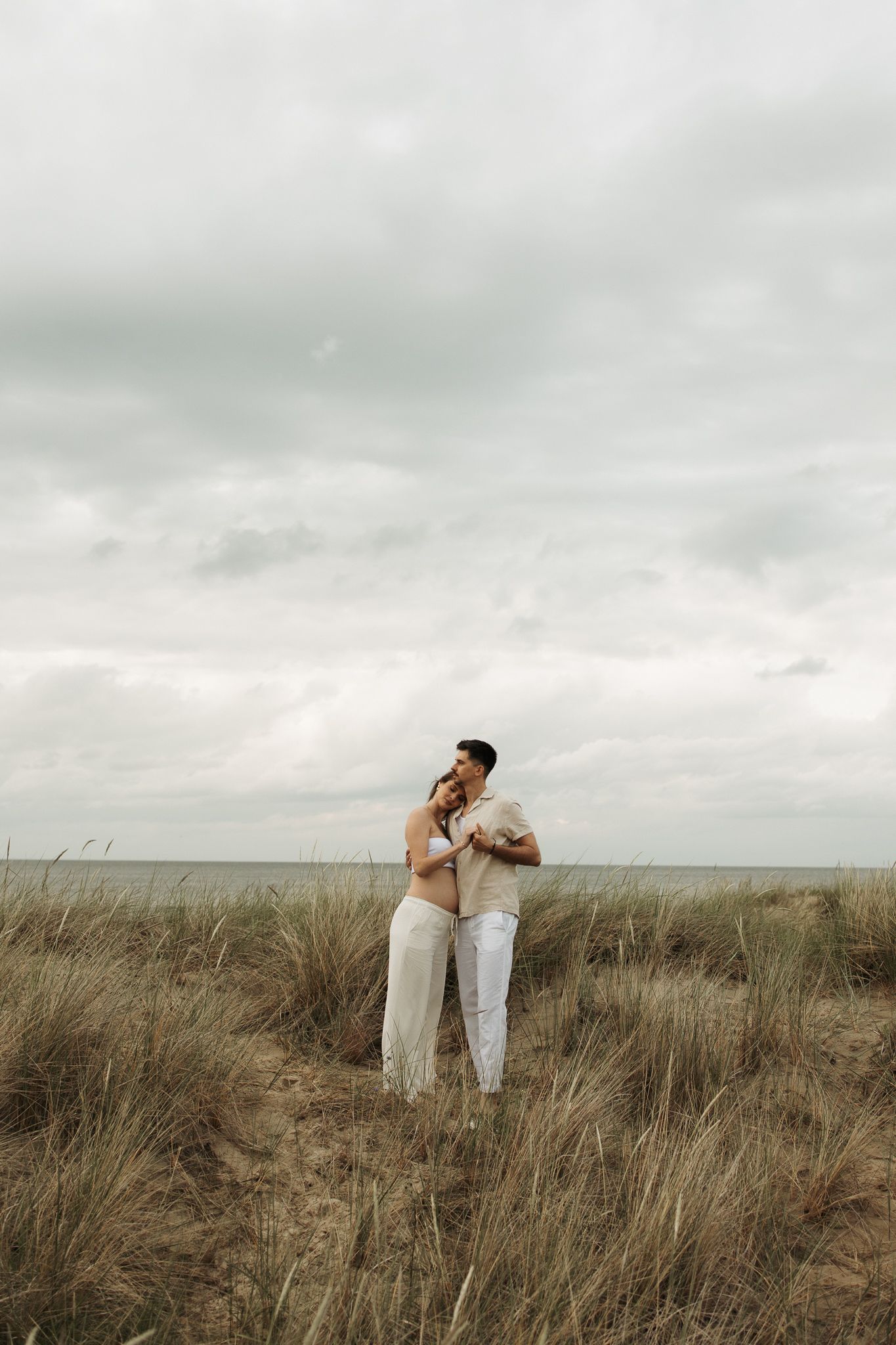 Expecting couple embracing in coastal grass dunes with ocean horizon under moody overcast sky.