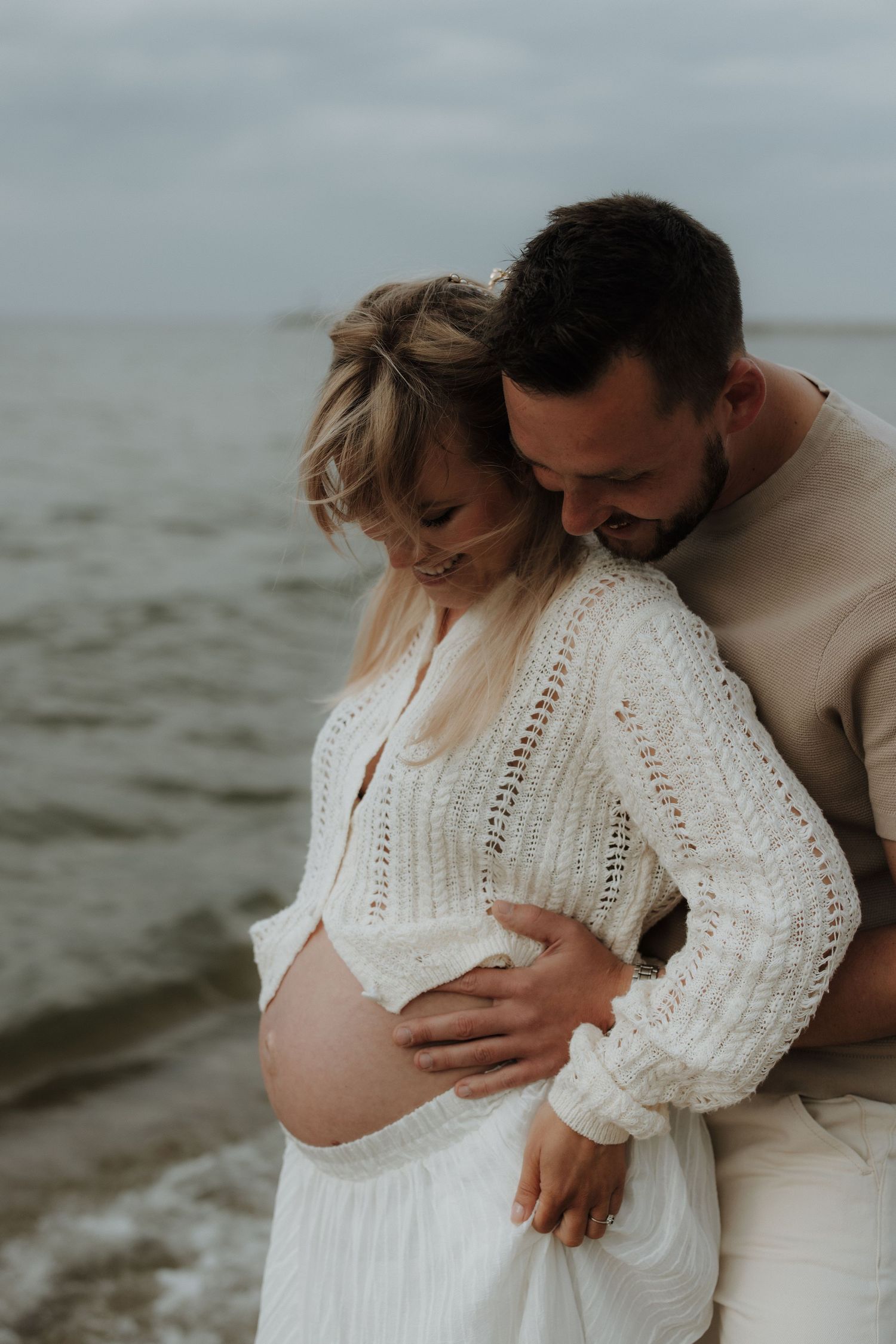 Couple embracing on beach, pregnant woman in white knit dress, man in beige sweater, ocean backdrop.