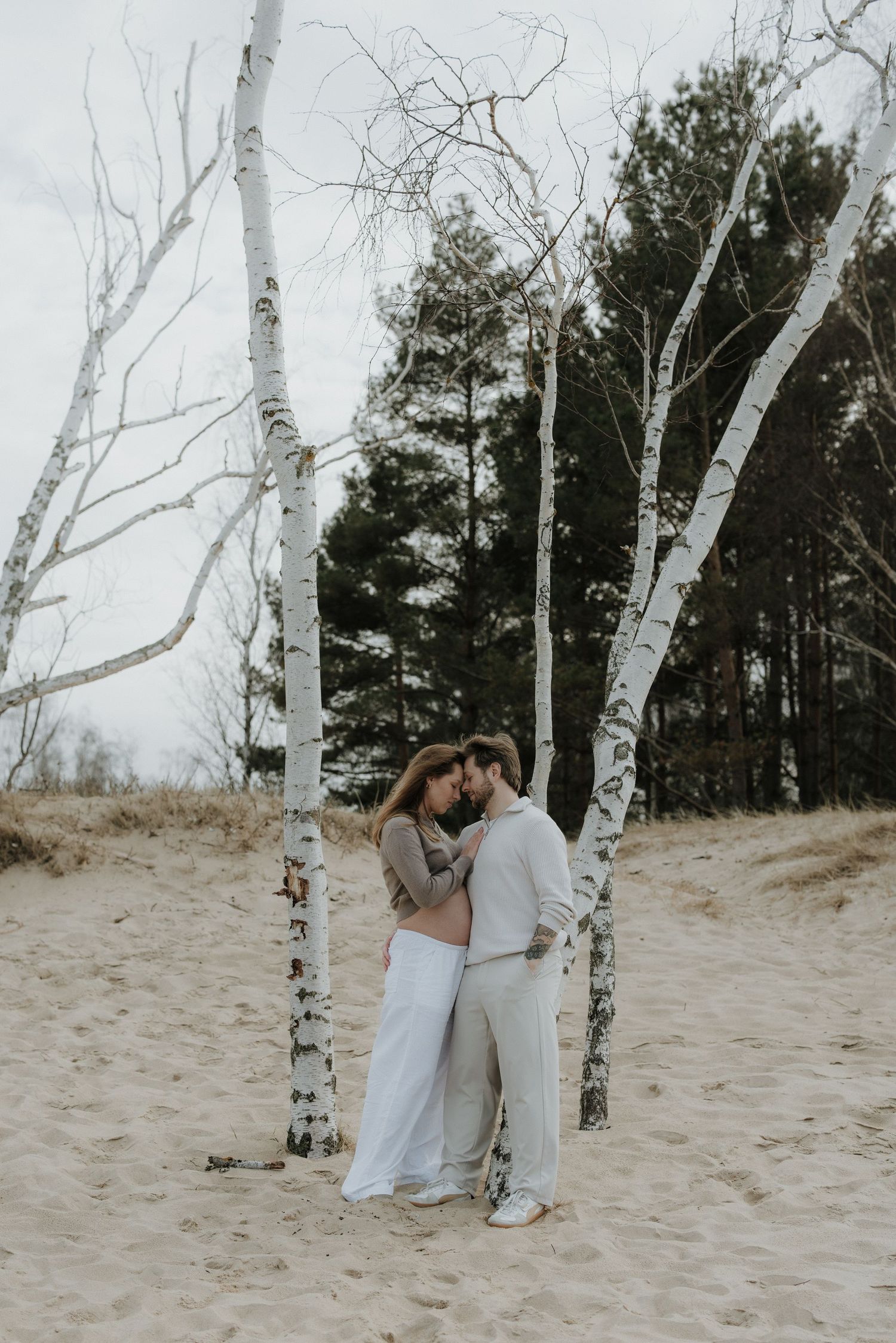 Couple embracing between white birch trees on sandy beach with evergreen forest backdrop.