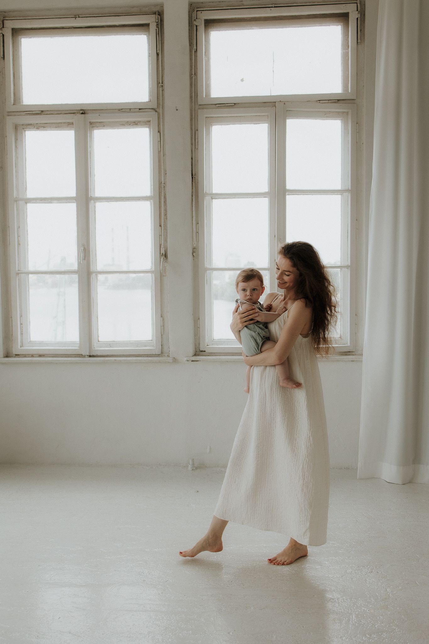 A barefoot mother in a white dress holds her baby in a bright white studio room.
