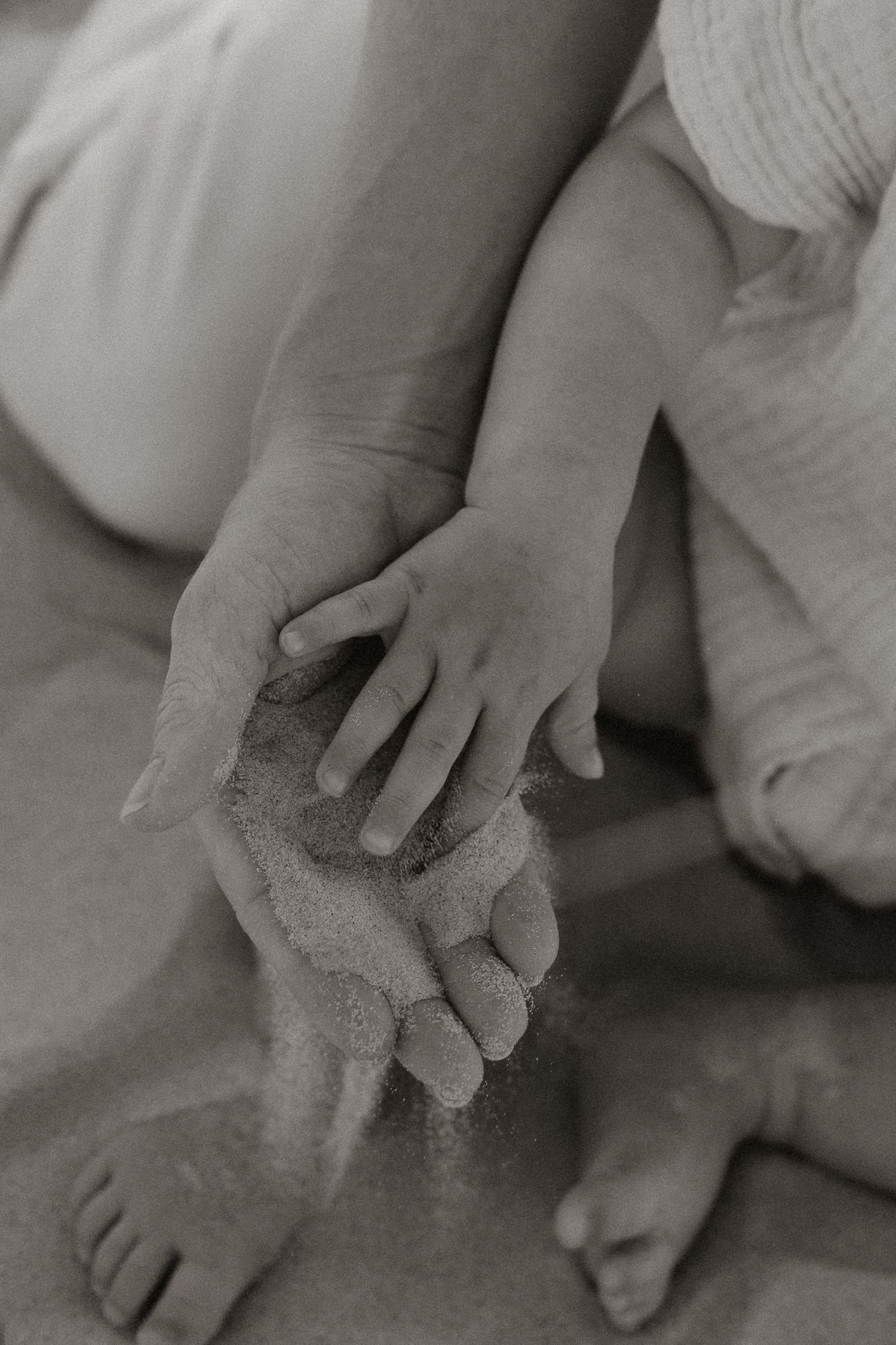 Black and white photo of adult and baby hands touching sand together at the beach.