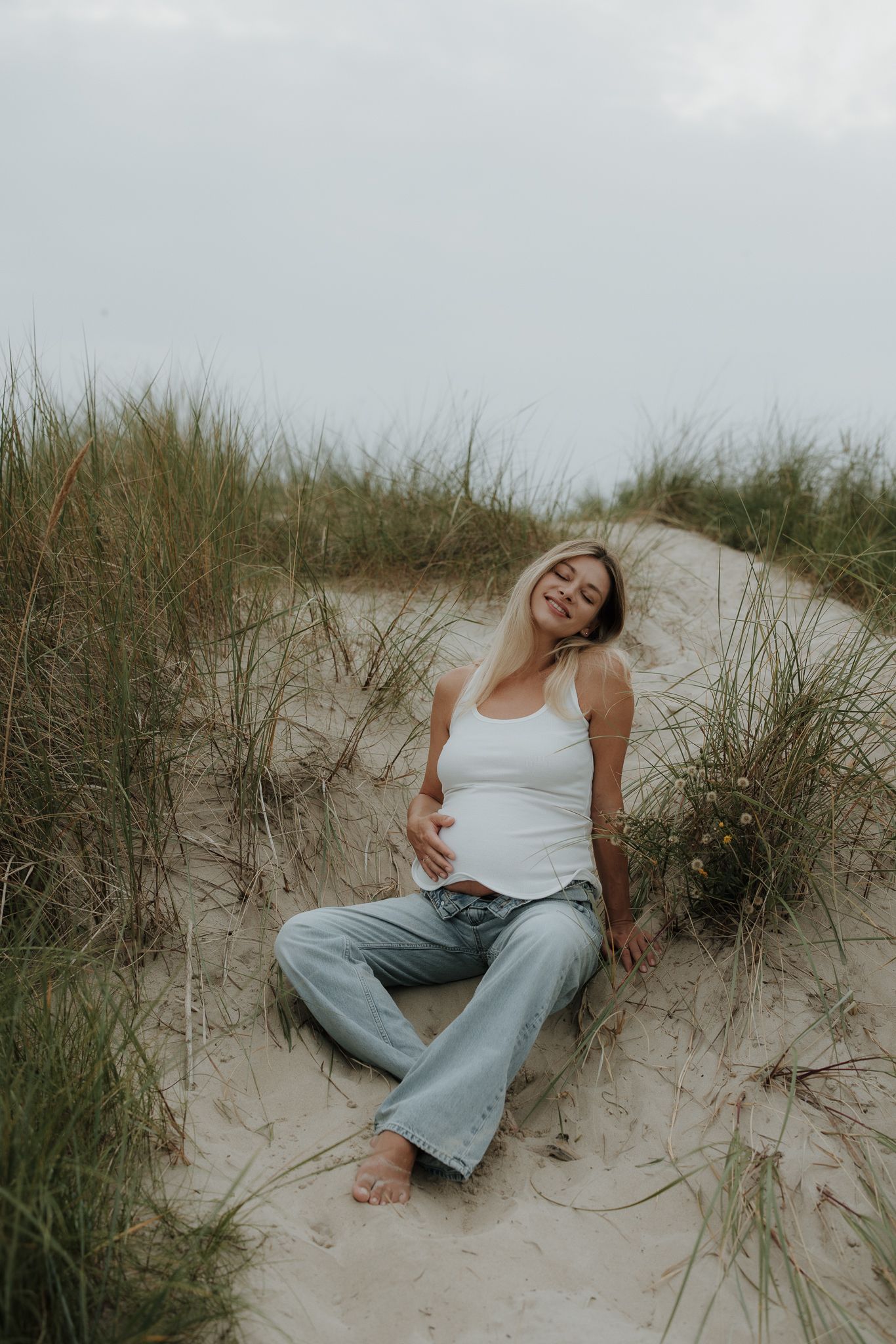 Smiling pregnant woman in white tank and jeans sitting relaxed in sandy beach dunes.