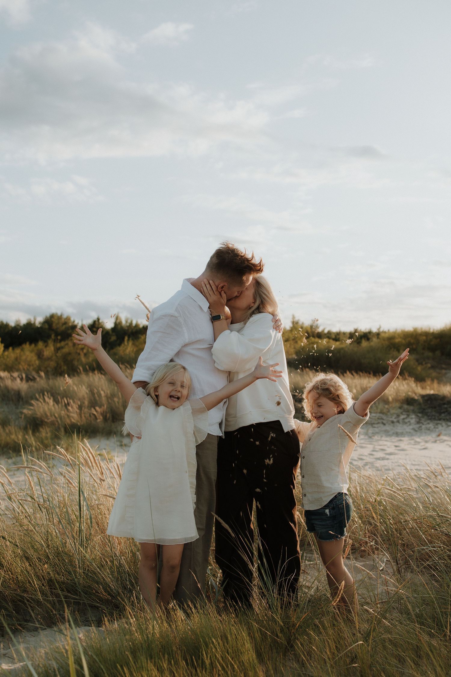 Happy family with parents embracing and two children raising arms in beach grass at dusk.