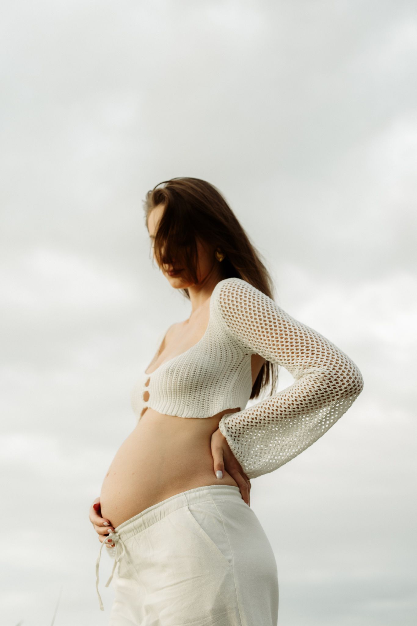 Pregnant woman in white mesh crop top and white pants holding her baby bump against an overcast sky.