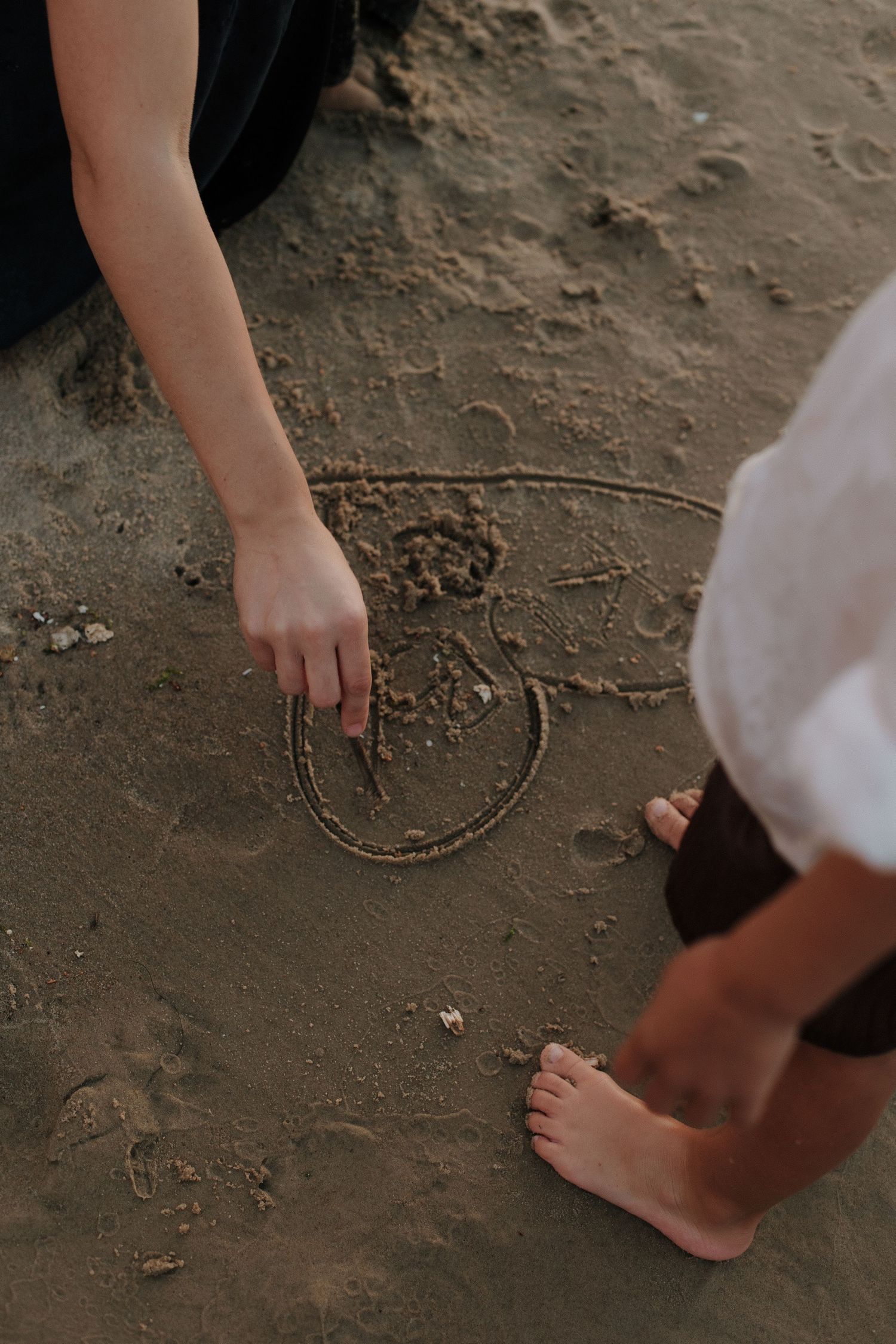 Person drawing a smiley face in wet sand at the beach with bare feet visible nearby.