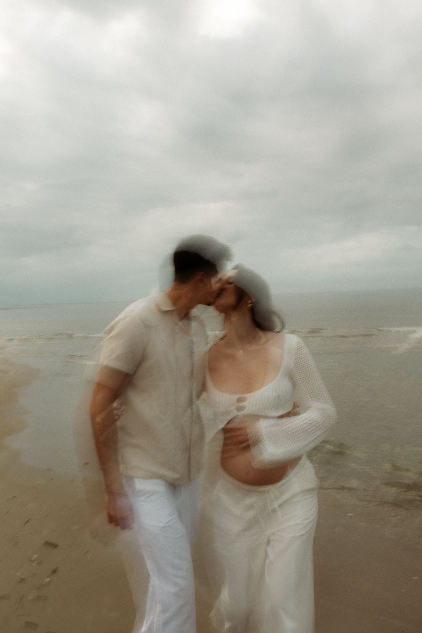 Pregnant couple sharing a tender kiss on a windy beach with overcast skies.