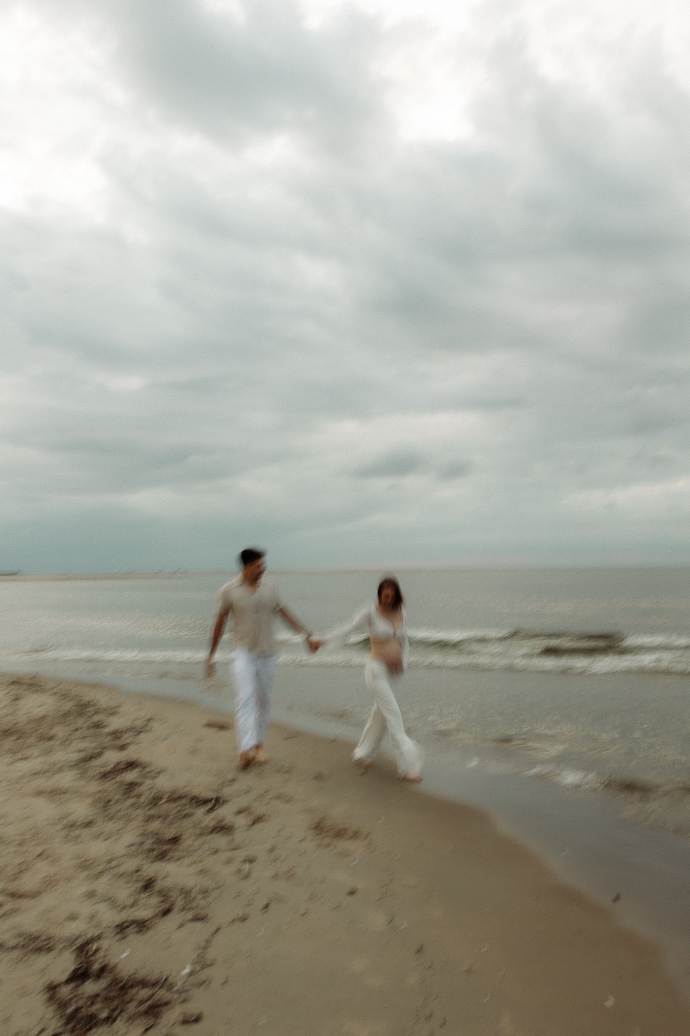 Couple holds hands walking along shoreline with dramatic cloudy sky in motion blur.