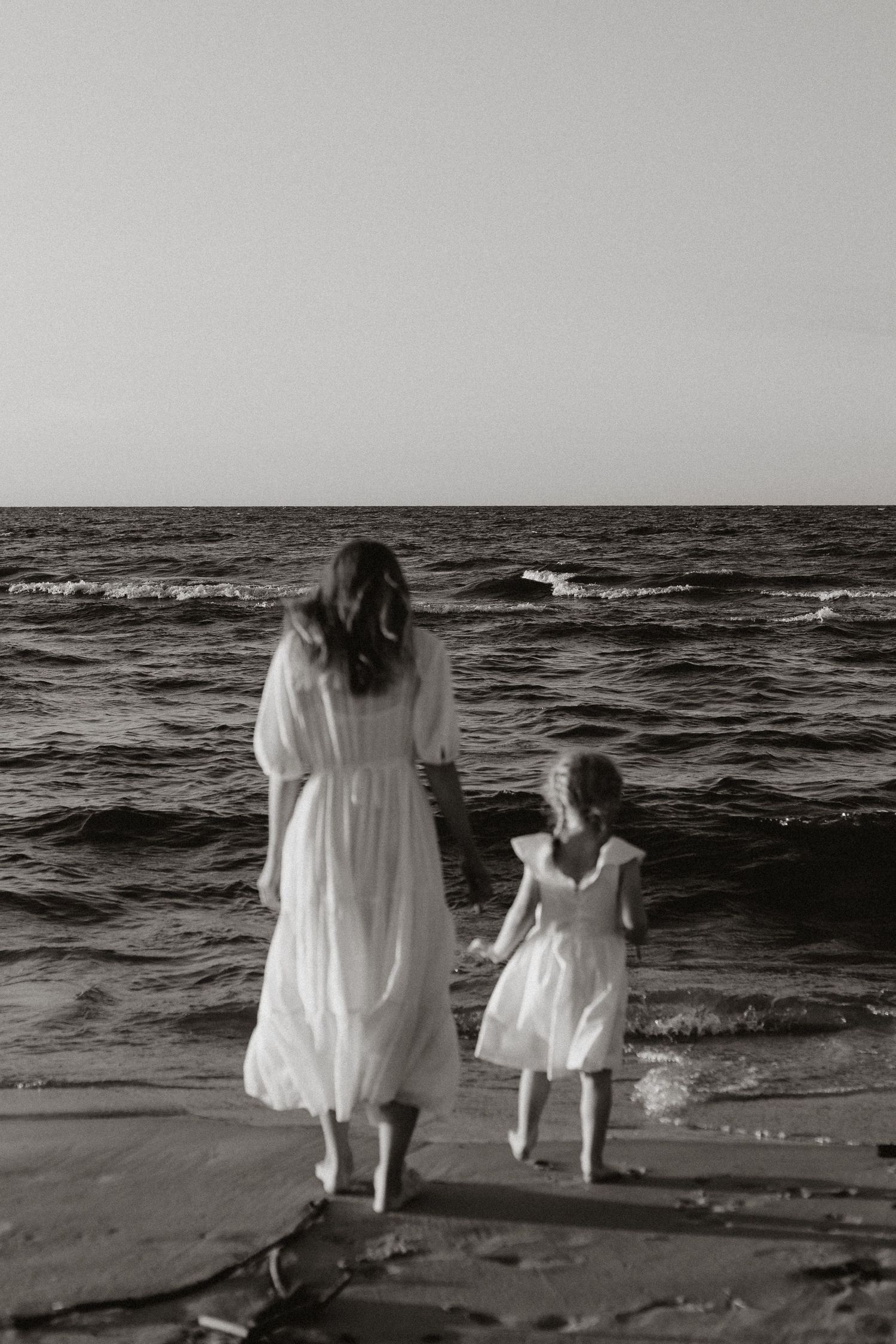 Black and white photo of mother and daughter in white dresses walking toward ocean waves.