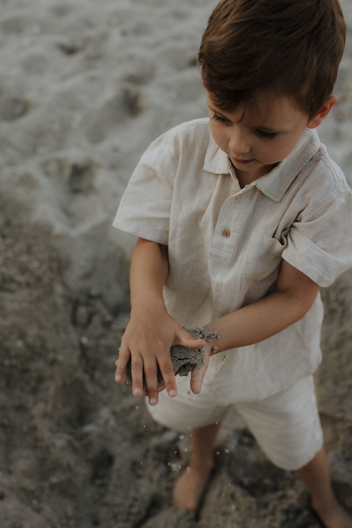 Young boy in white linen outfit playing with sand and rocks on a beach.