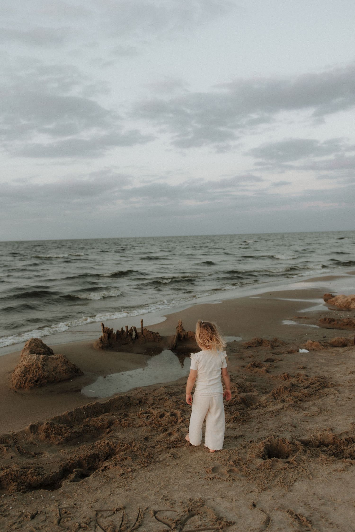 Young child in white outfit standing before ocean waves on a cloudy beach day.