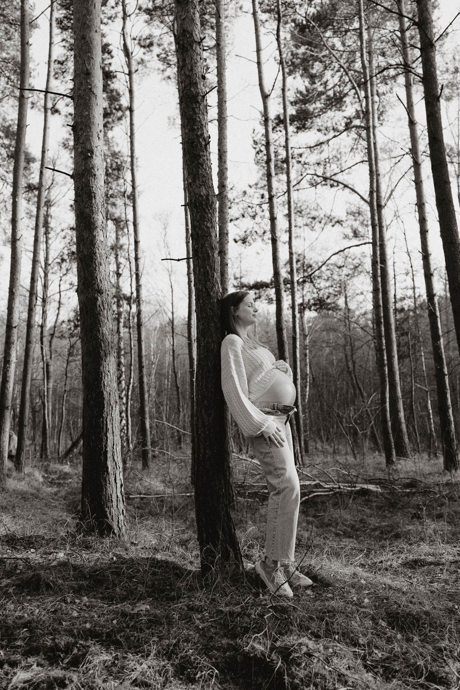 Black and white portrait of a pregnant woman leaning against a pine tree in a forest.