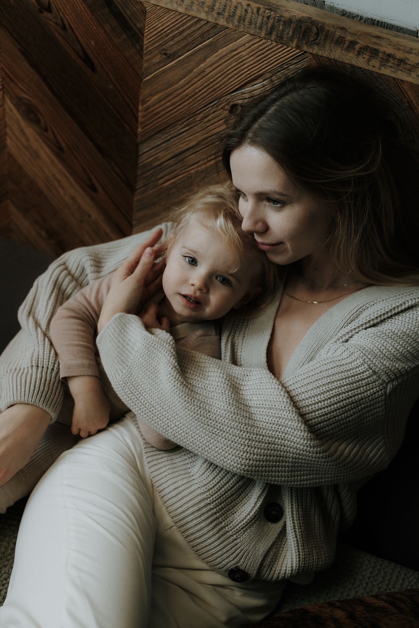 Mother in beige knit cardigan gently embracing her toddler against wooden wall.