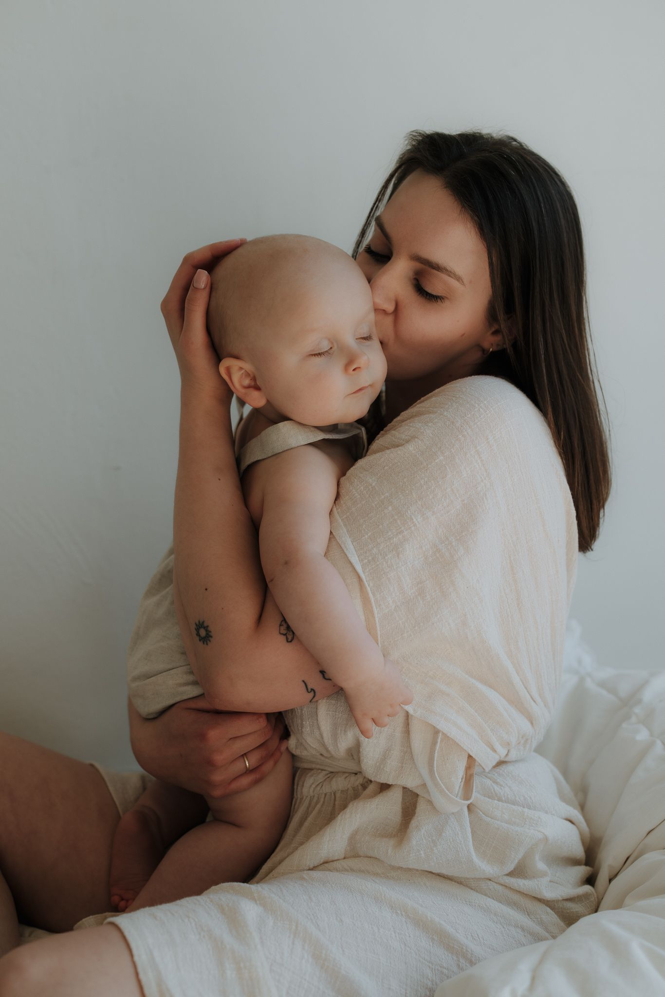 Mother in cream top tenderly kissing her sleeping baby on the head while seated.