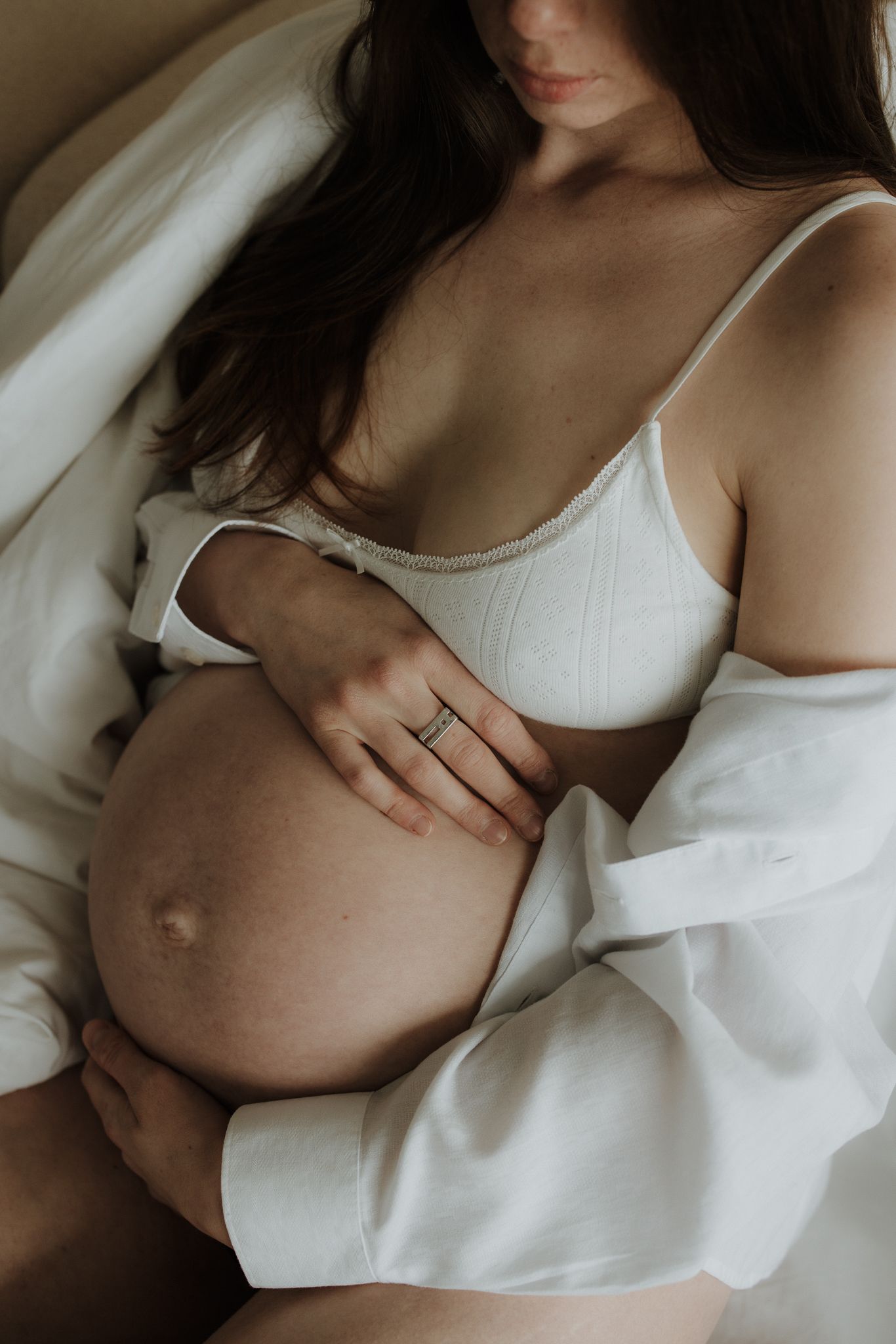 Pregnant woman in white lingerie and open robe gently holding her baby bump.