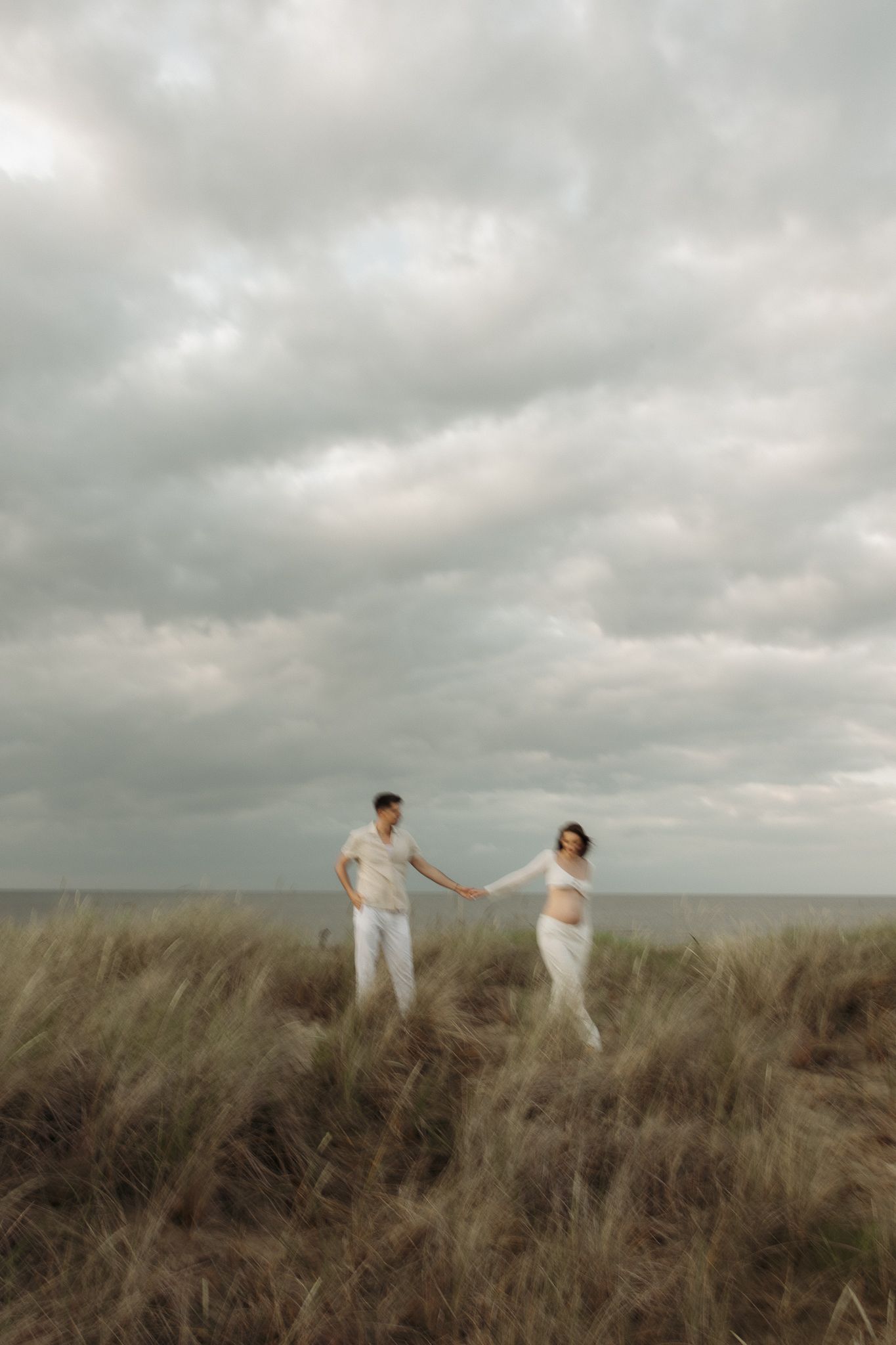 Expecting couple holding hands on a grassy coastal dune with the ocean in the background.