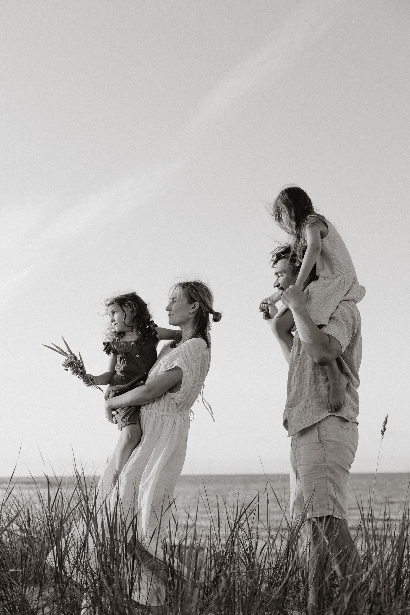 Black and white beach photo of a family of four, parents carrying children on a sunny day.