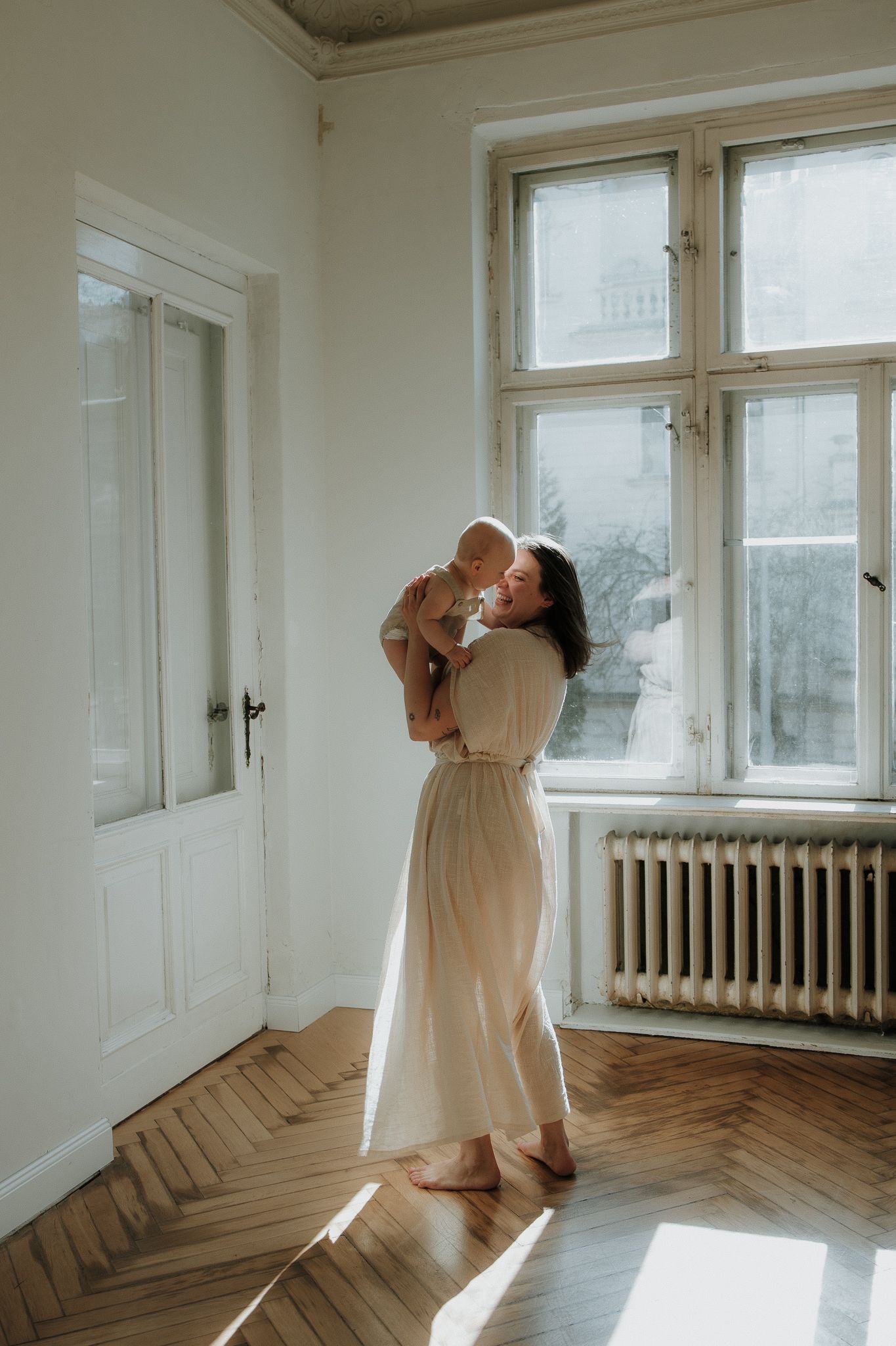 Mother in cream dress holding baby up near sunlit window in bright white room.