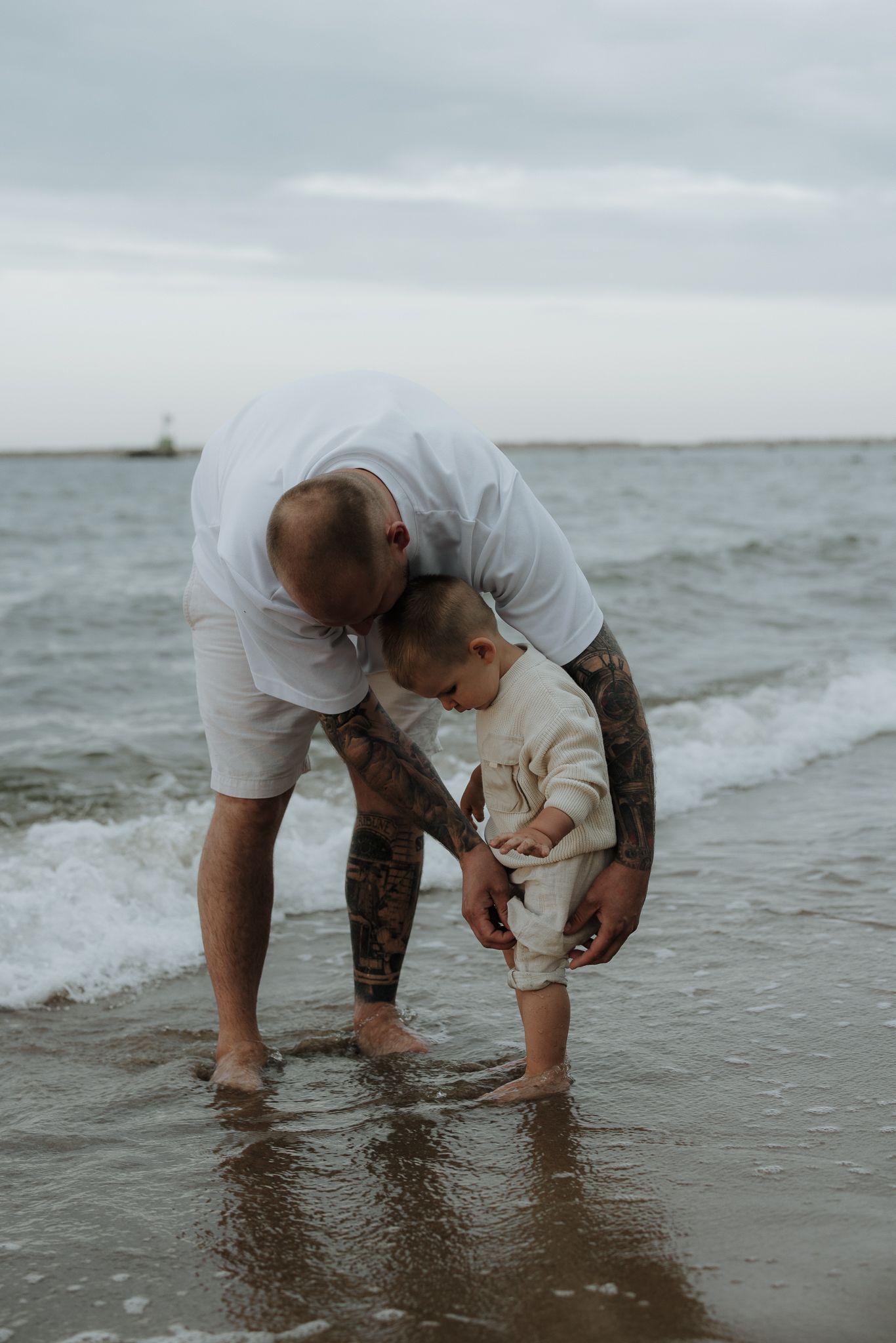 Tattooed father helping toddler stand at ocean shoreline on a cloudy day.