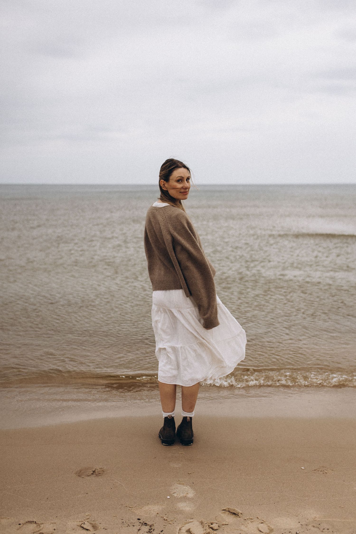 Woman in white skirt and brown sweater standing on sandy beach with calm ocean waves behind her.