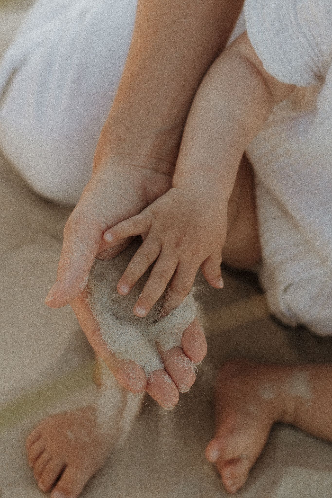 Mother and baby playing with sand together, letting it fall through their hands at the beach.