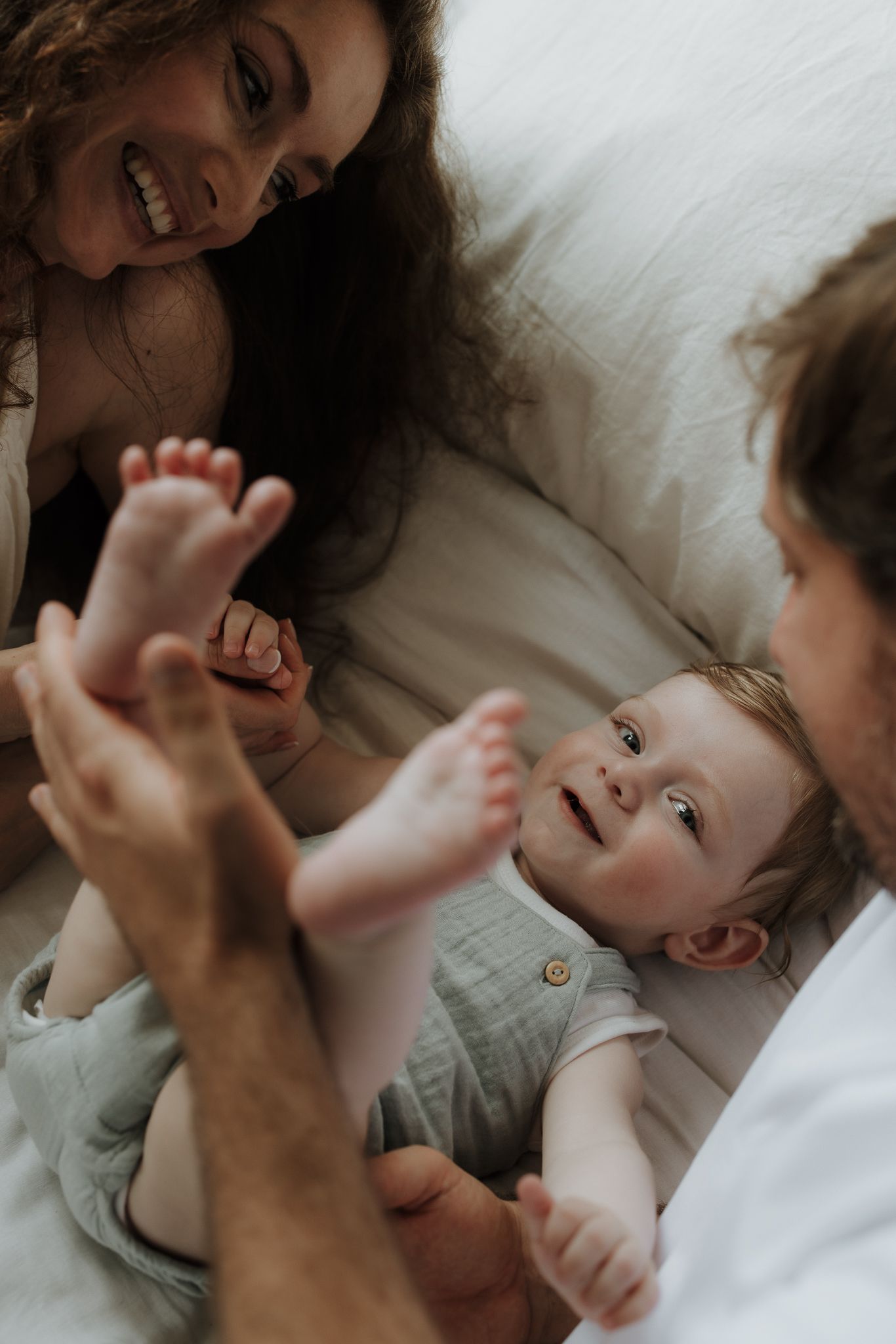 Happy parents playing with smiling baby lying on white bedding at home.