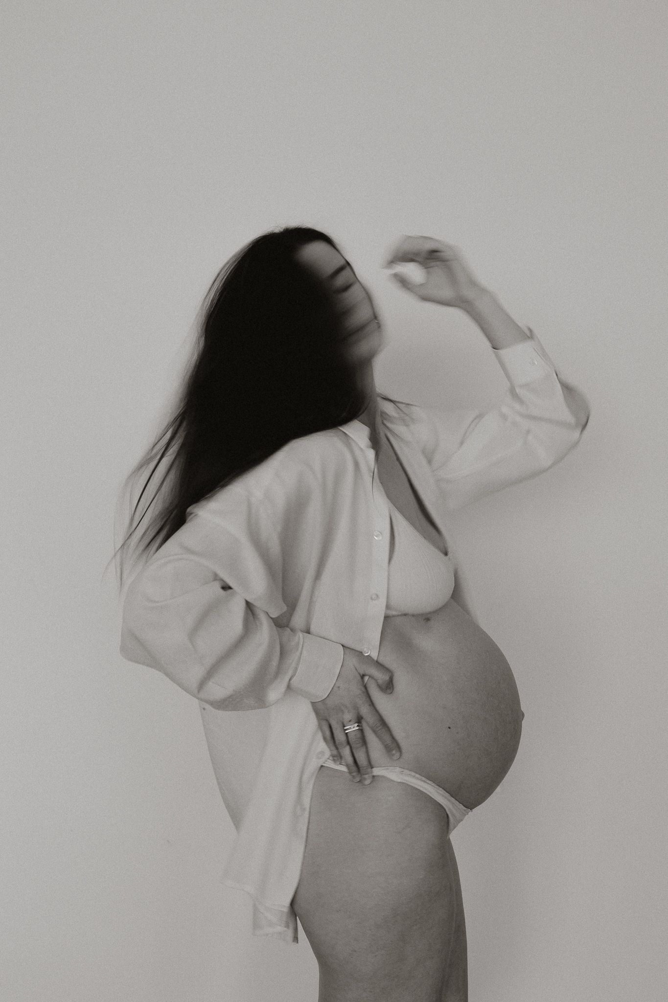 Black and white artistic photo of a pregnant woman in white lingerie with hair covering her face.