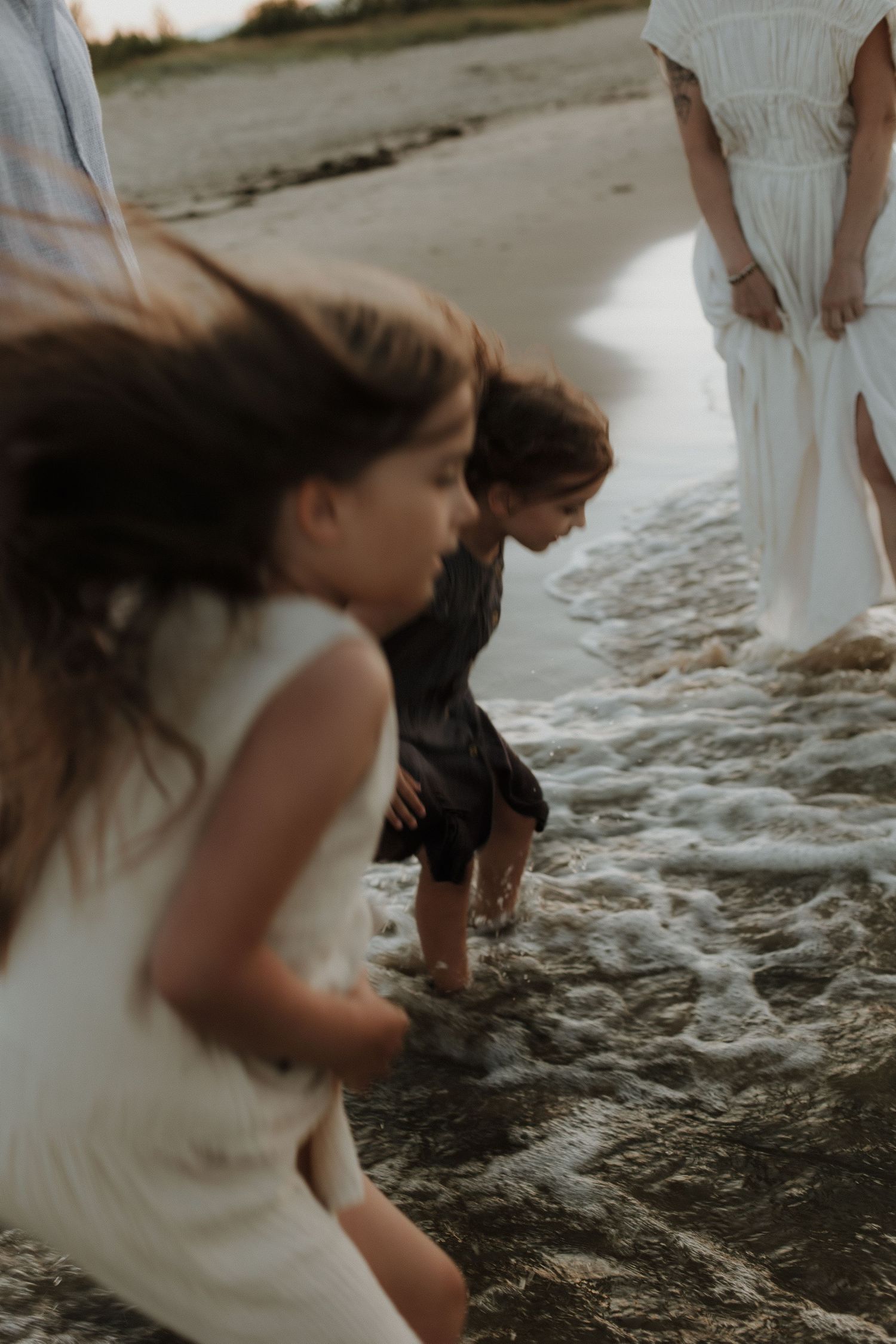Children playing in ocean waves at beach with women in white dresses nearby.