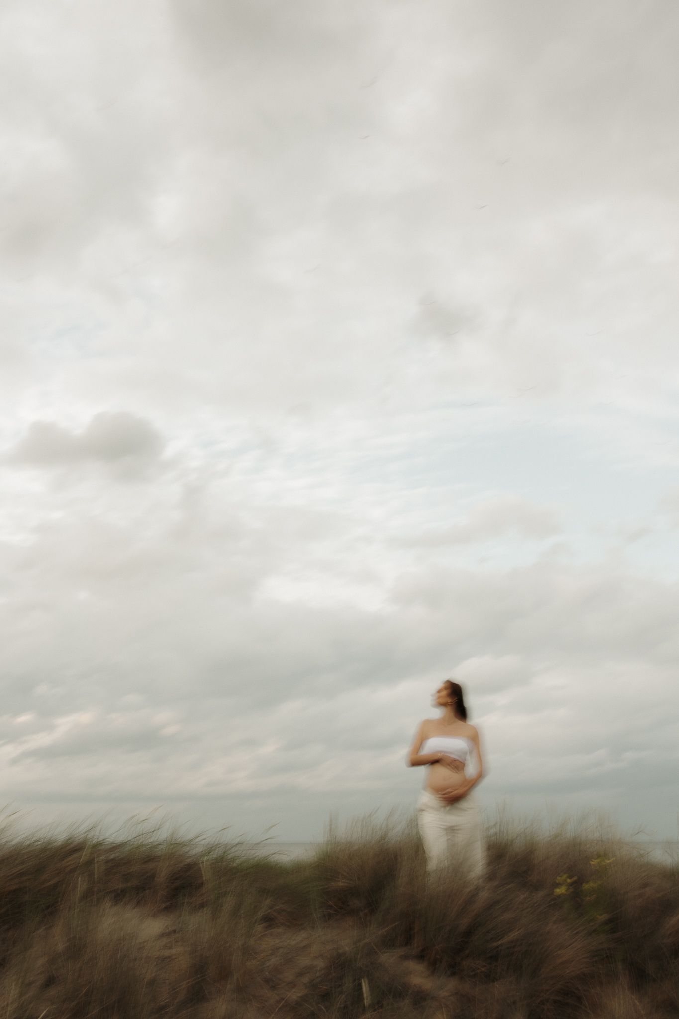 Pregnant woman in white standing on grassy dune under expansive cloudy sky.