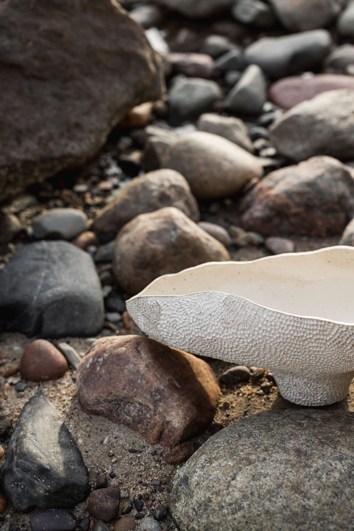 White textured ceramic bowl resting on smooth river rocks and pebbles on a rocky beach shoreline.