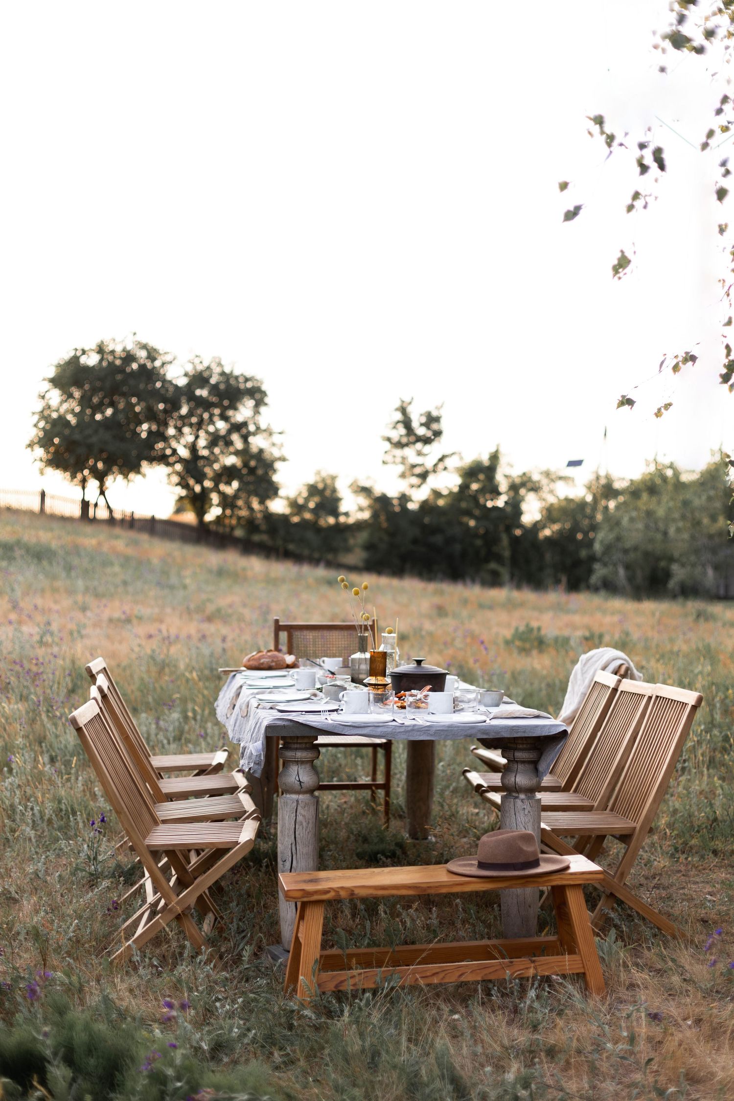 Rustic wooden outdoor dining table set in a meadow at golden hour, styled with glassware and a hat on the bench.