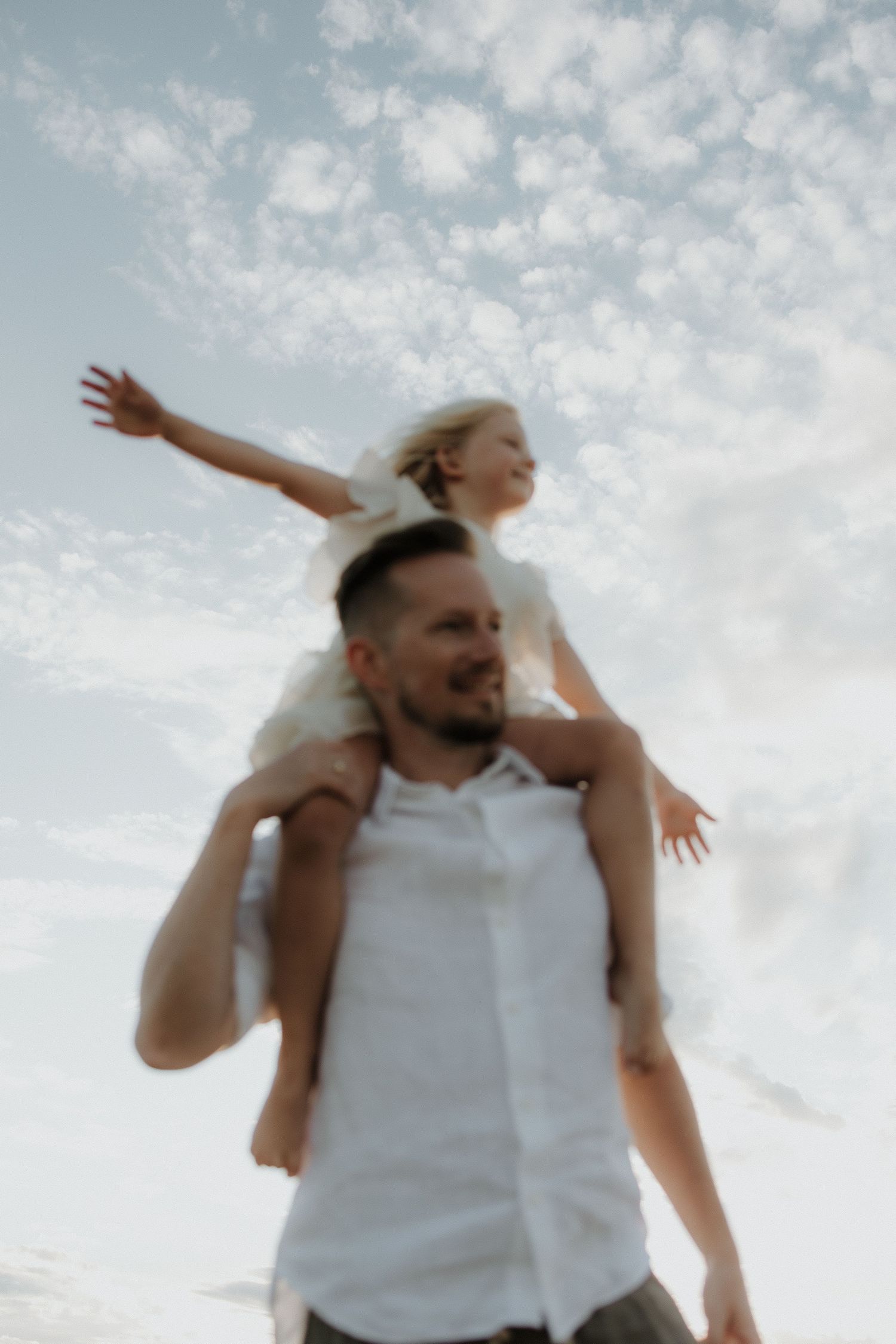 Joyful child with arms spread wide sitting on father's shoulders against a cloudy sky.