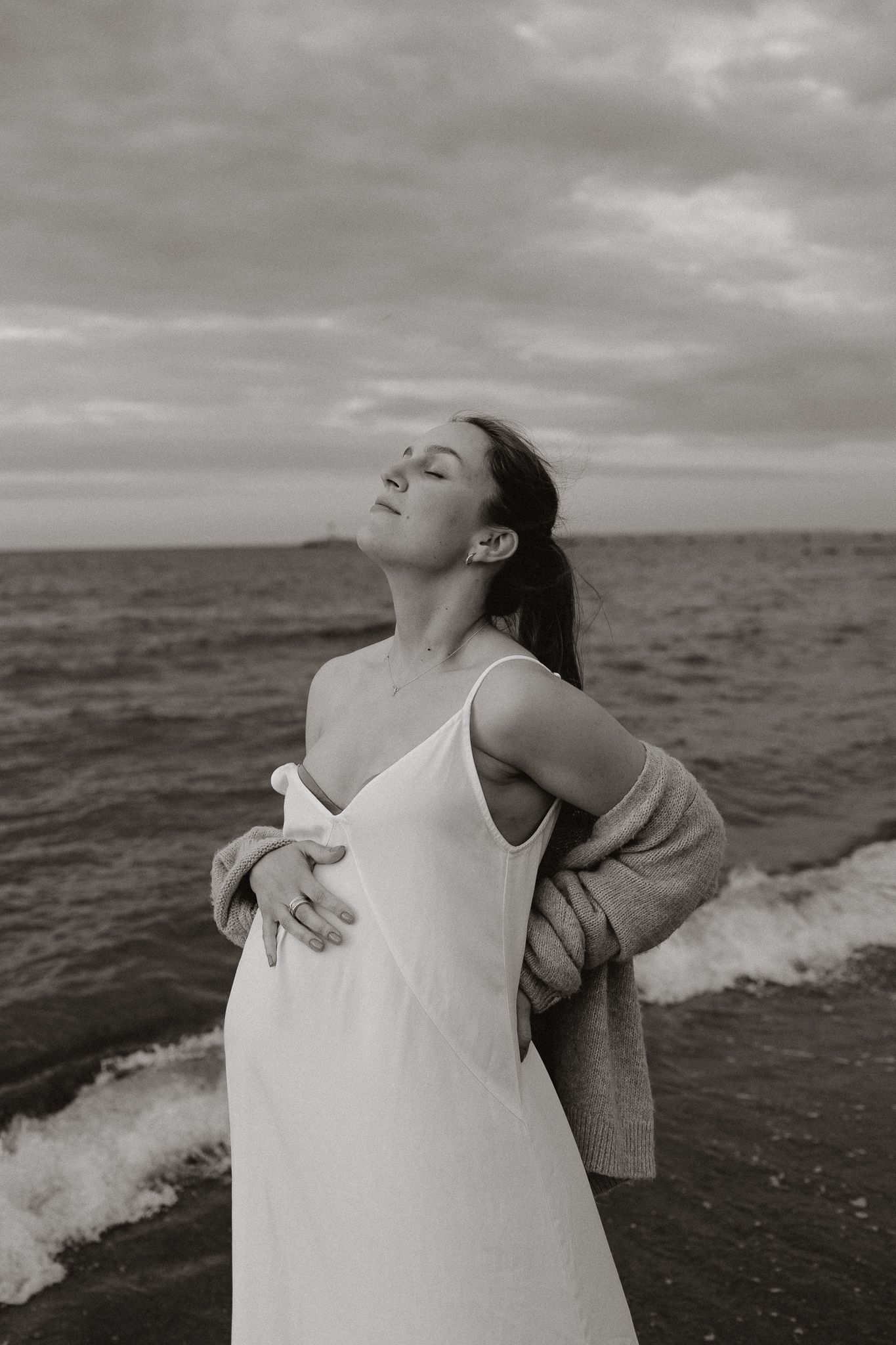 Black and white portrait of pregnant woman in white dress holding belly near ocean shore.