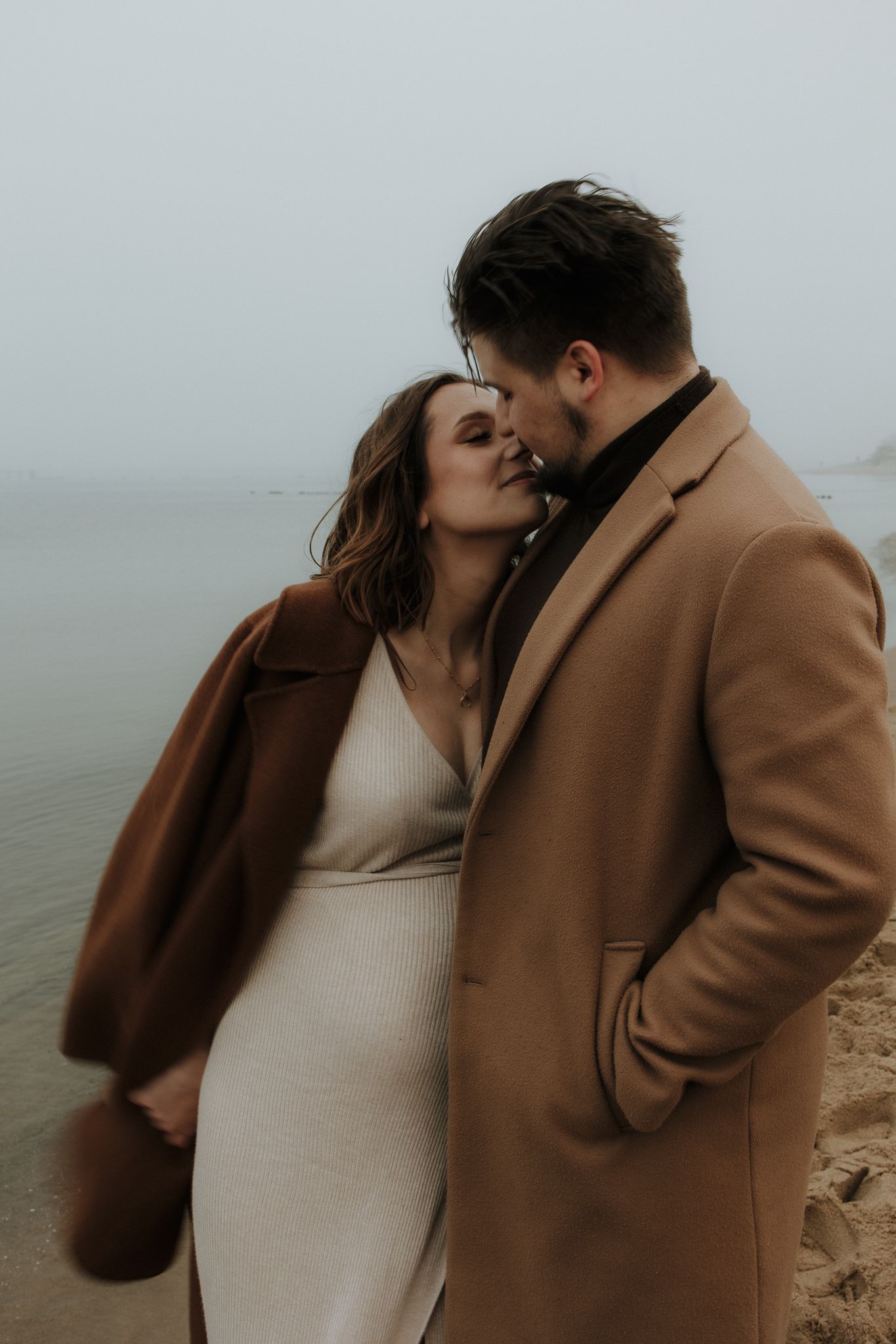 Couple sharing a kiss on foggy beach, woman in beige dress and man in camel coat.