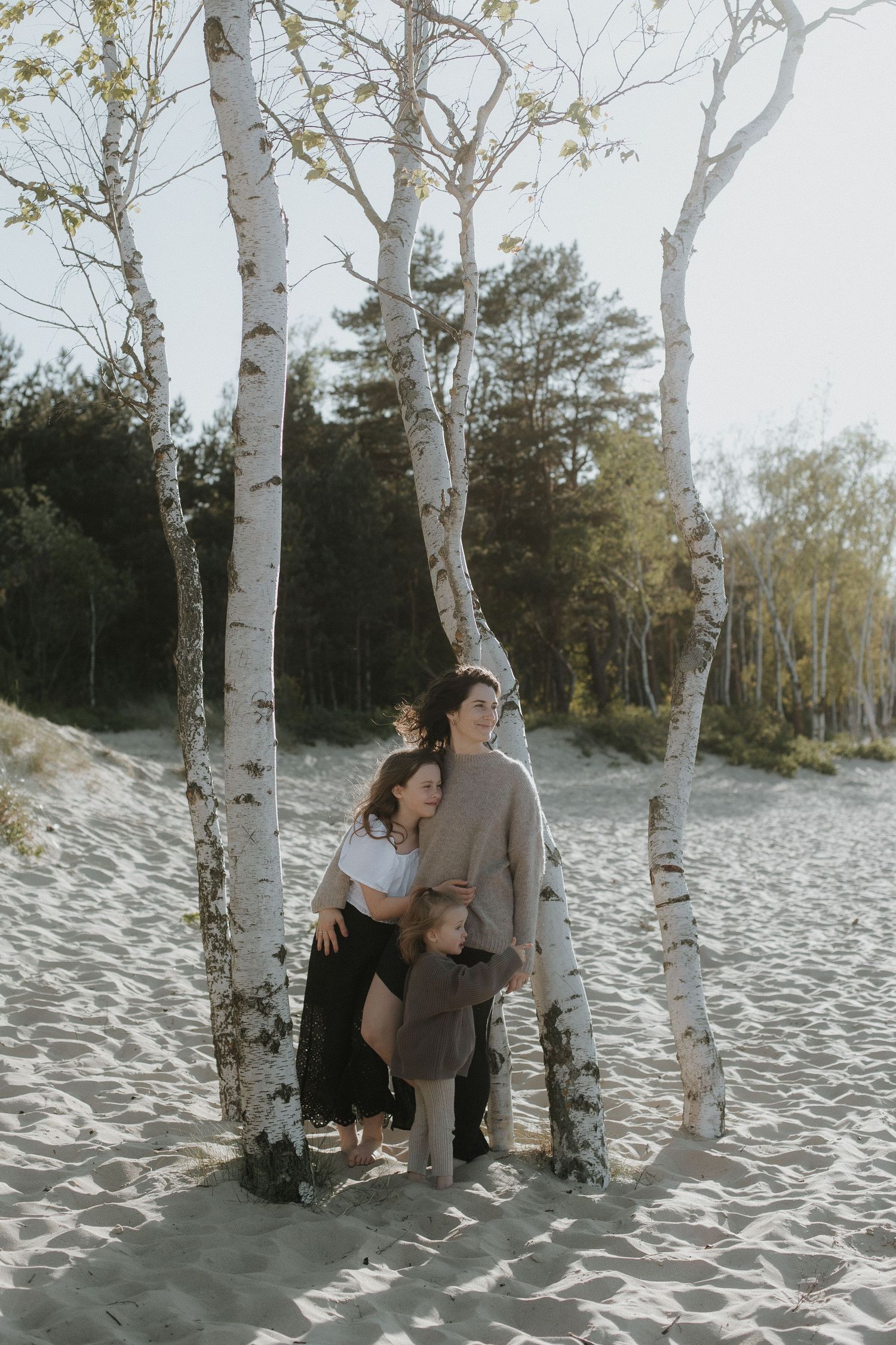 Mother and two children embracing among white birch trees on sandy dunes in soft sunlight.