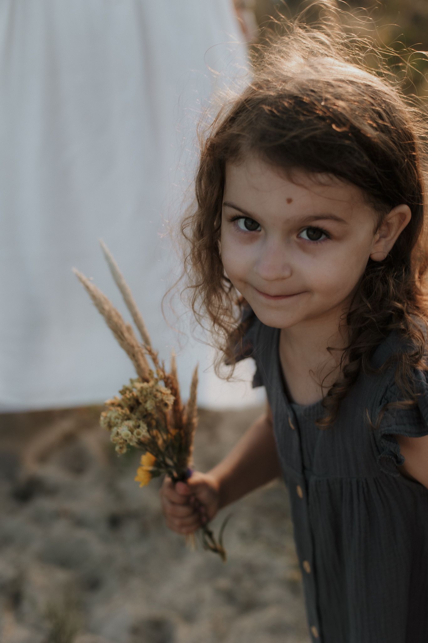 Curly-haired little girl in gray dress holding wildflowers and smiling at camera.