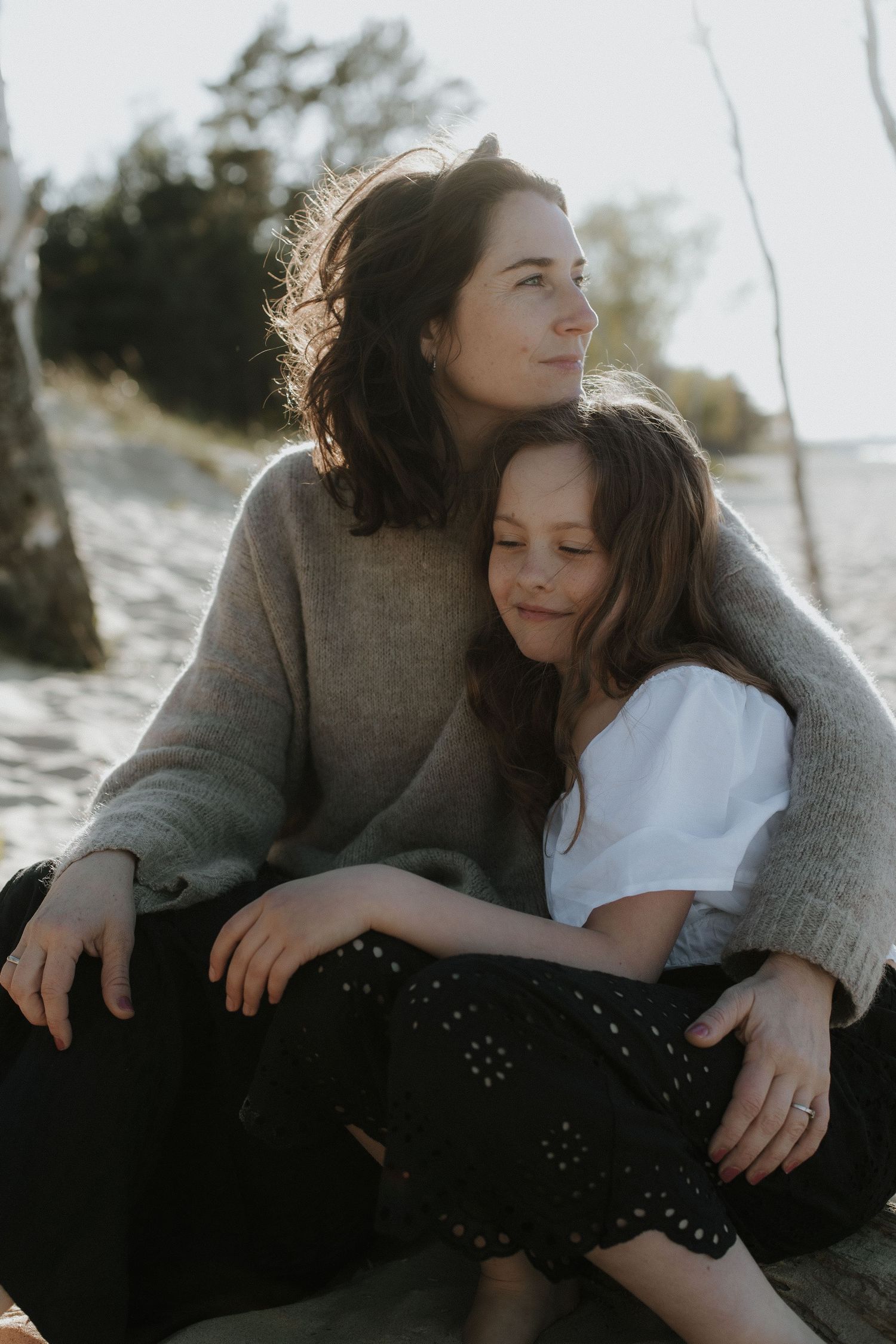 Brunette mother in gray sweater embracing her daughter while sitting on a sunny beach together.