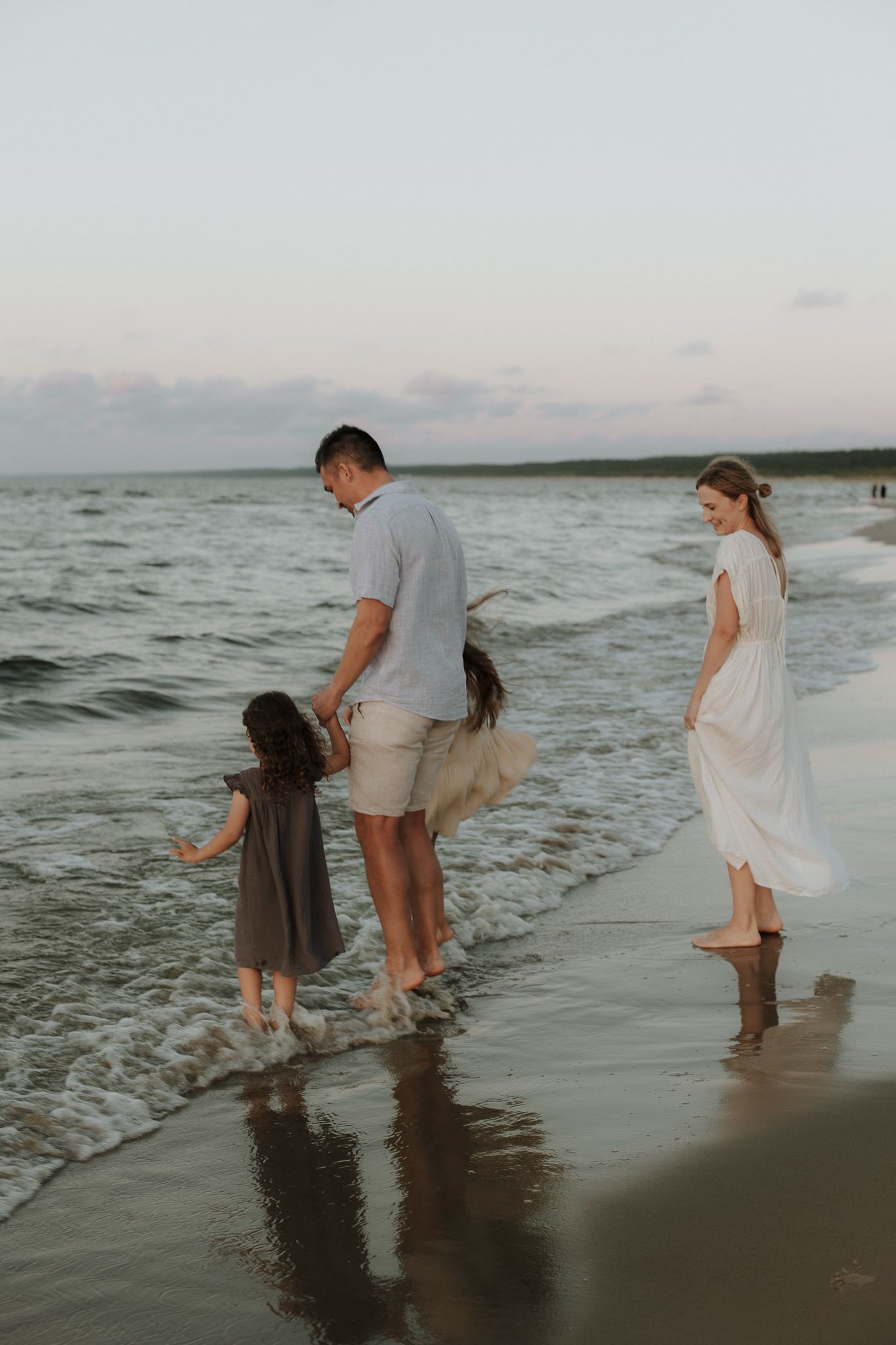Family of four walking barefoot along a sandy beach shore at sunset, parents and two children enjoying ocean waves.
