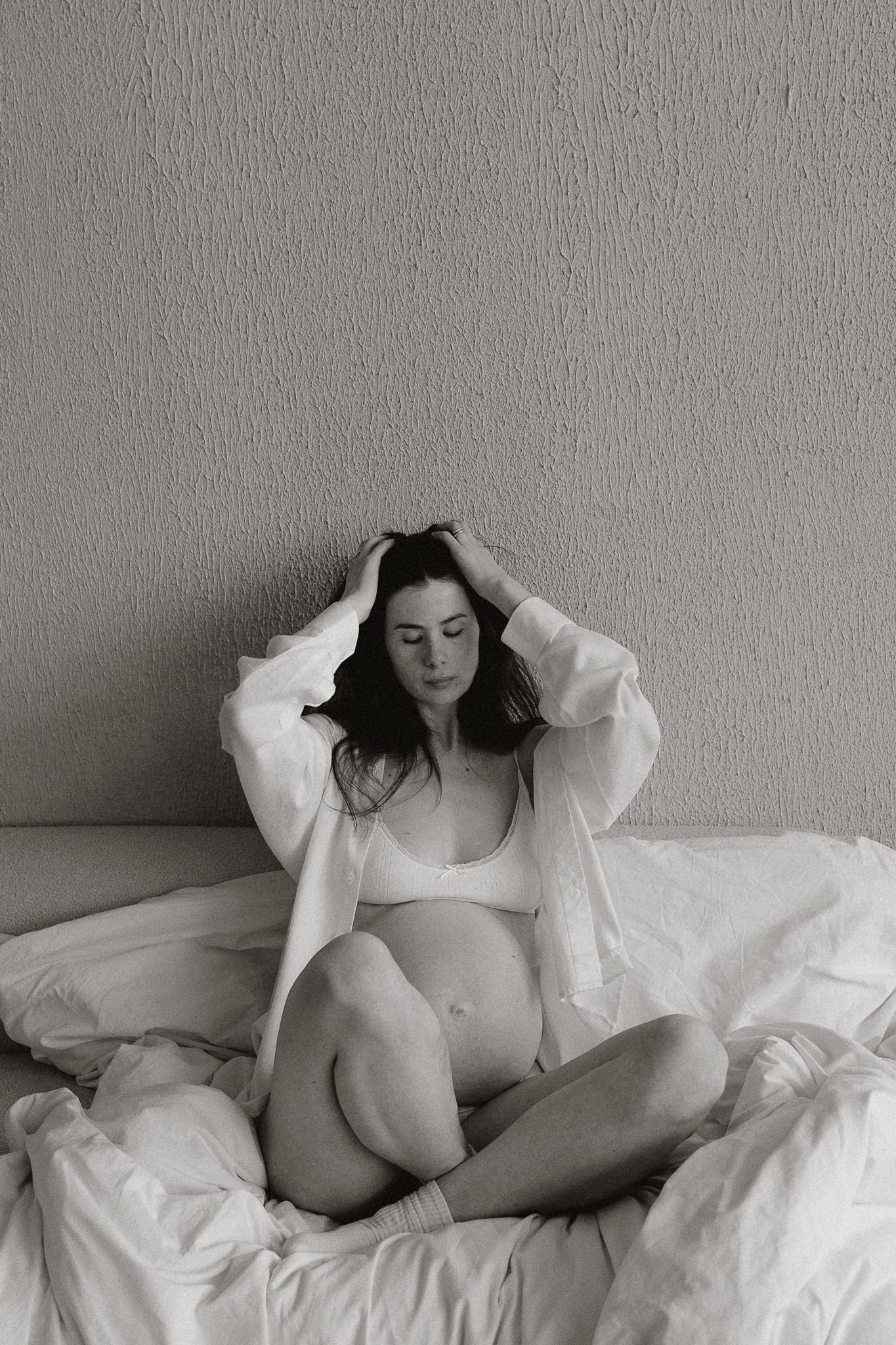 Black and white photo of a pregnant woman in white lingerie sitting cross-legged on a bed with hands in hair.