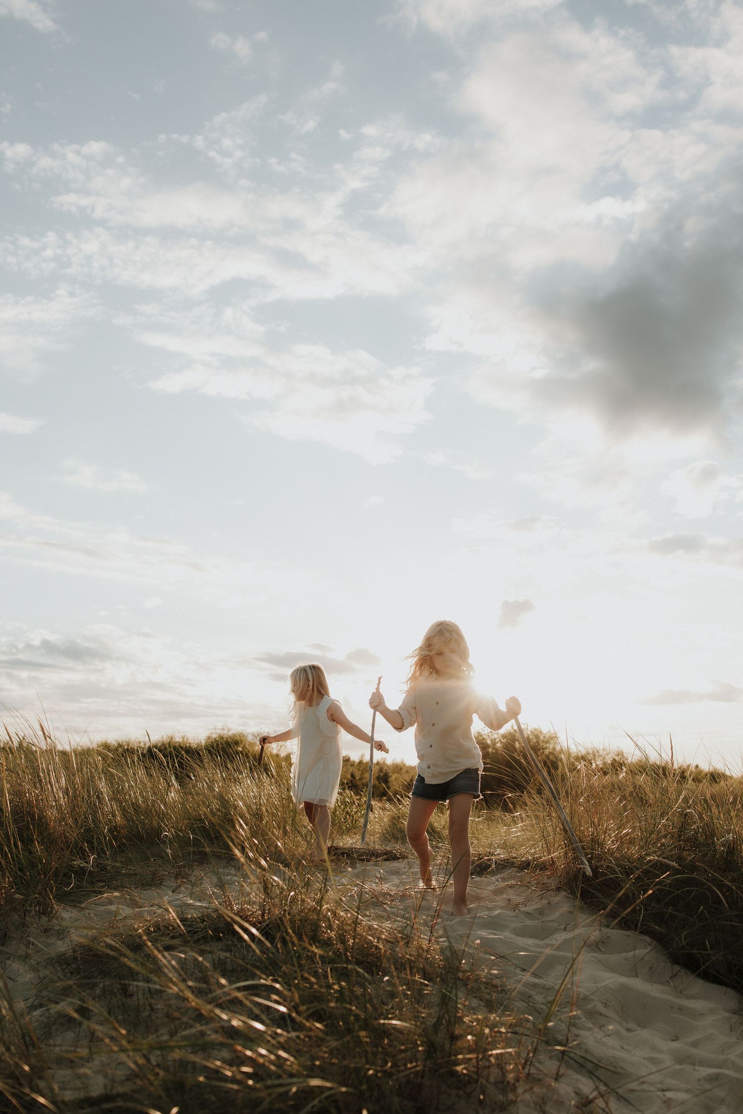 Two children walking on a sandy beach path through golden grass at sunset.