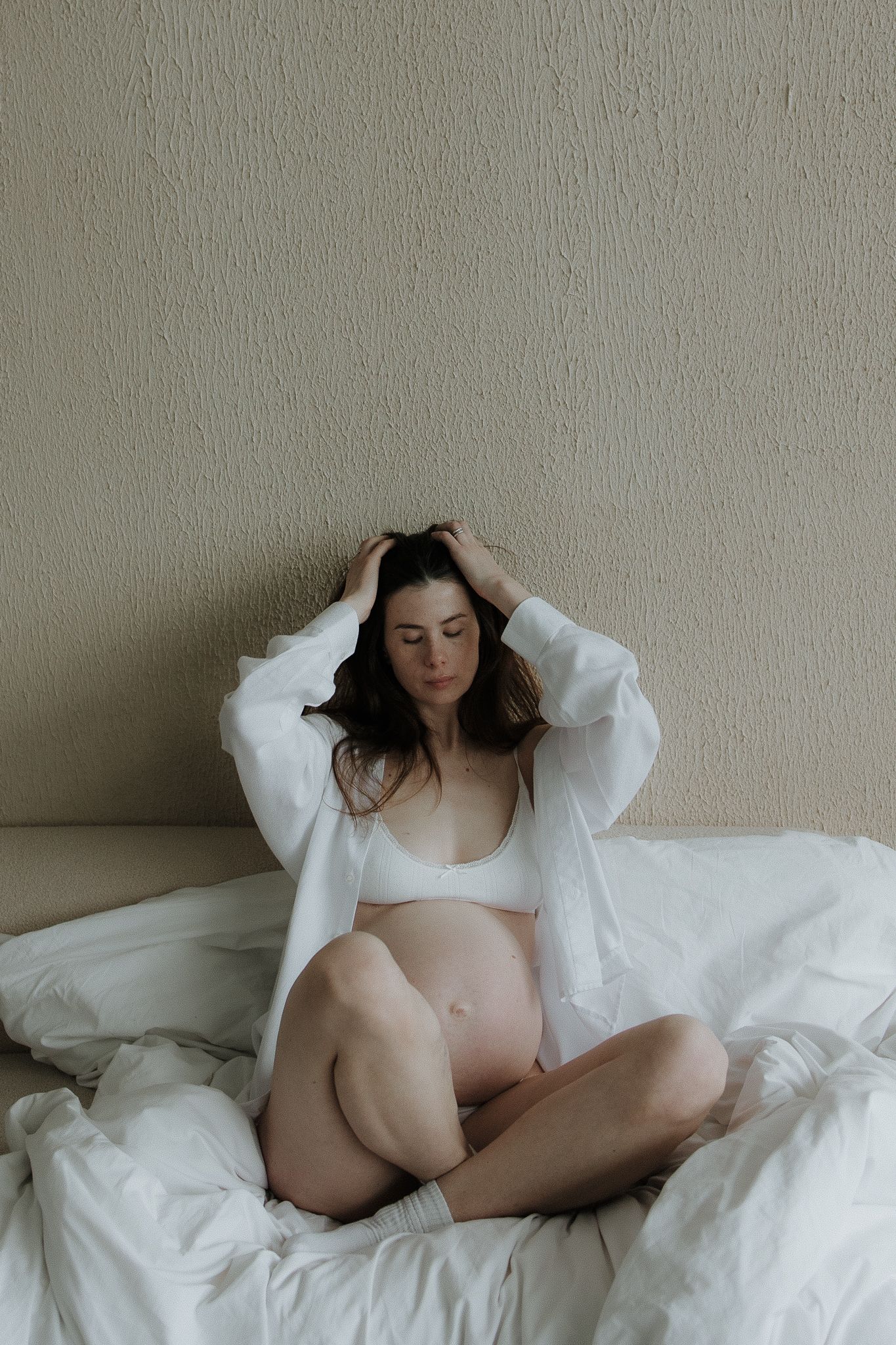 Pregnant woman in white shirt and lingerie sits cross-legged on bed holding her hair.