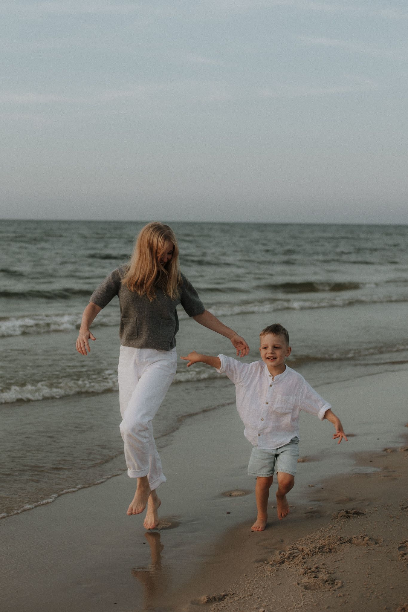 Mother and young son running barefoot along ocean shoreline at sunset on sandy beach.