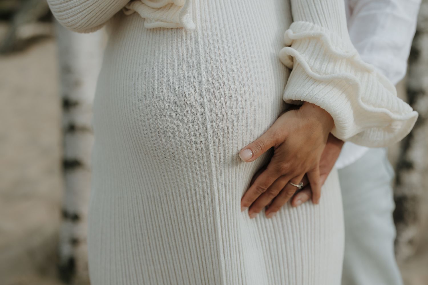 Close-up of pregnant belly in white ribbed dress with partner's hand gently resting on it.