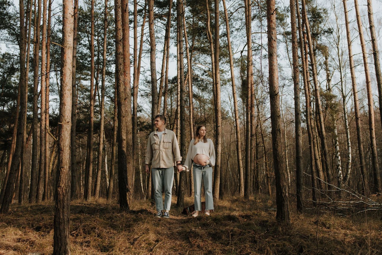 Couple holding hands while walking through tall pine forest in casual maternity photo session.