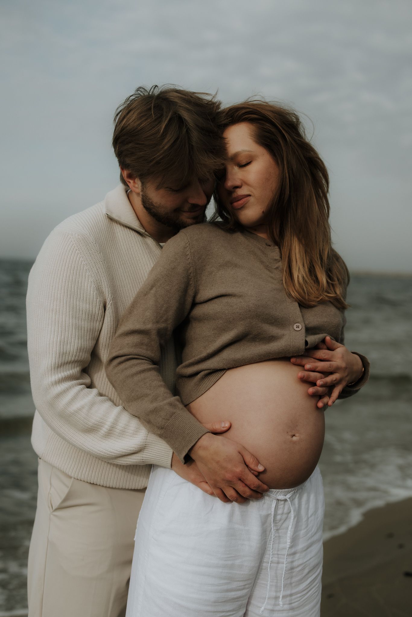 Man embracing pregnant woman from behind on overcast beach, hands on baby bump.