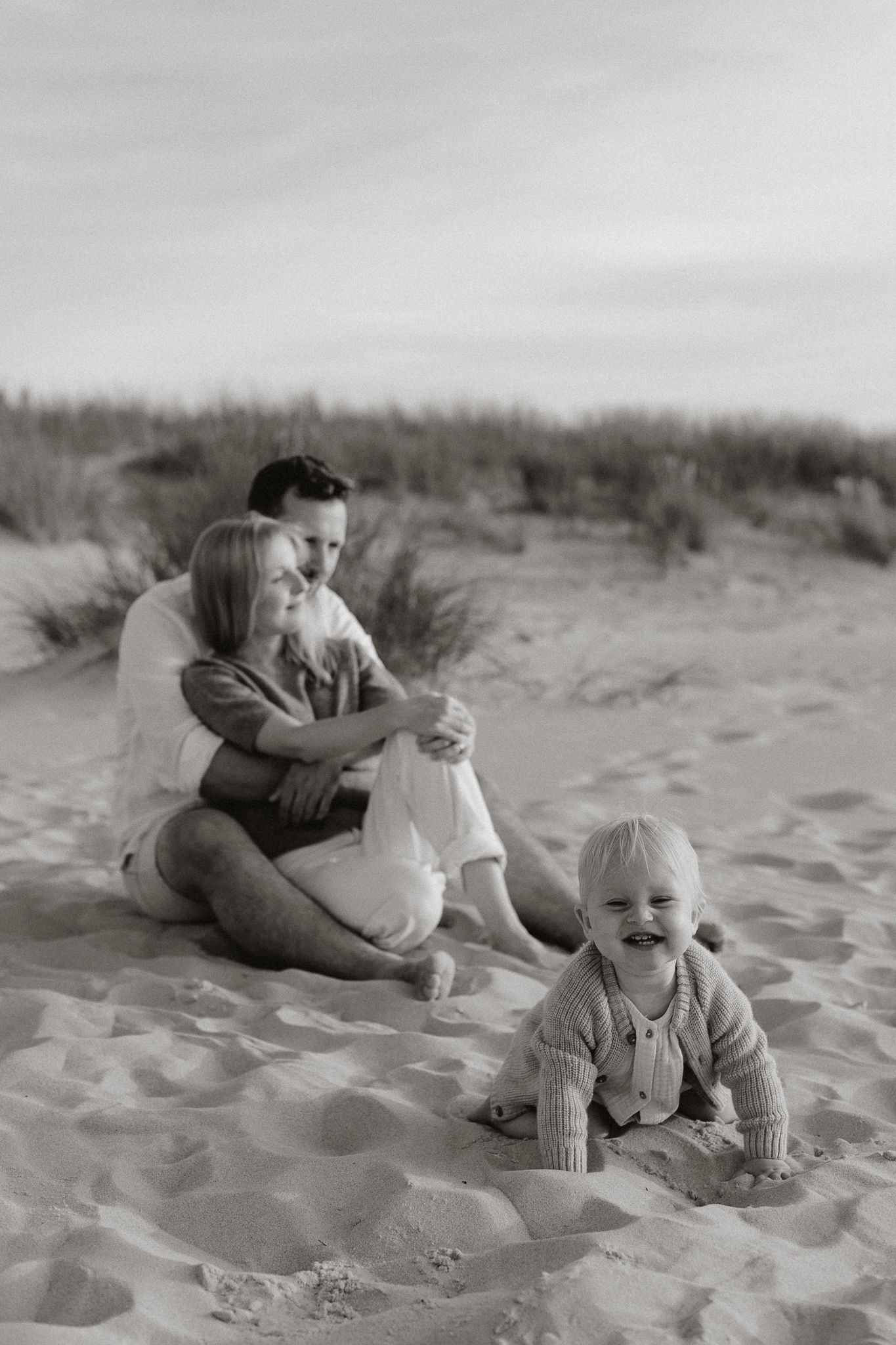 Black and white photo of parents sitting in sand dunes while smiling baby crawls nearby.
